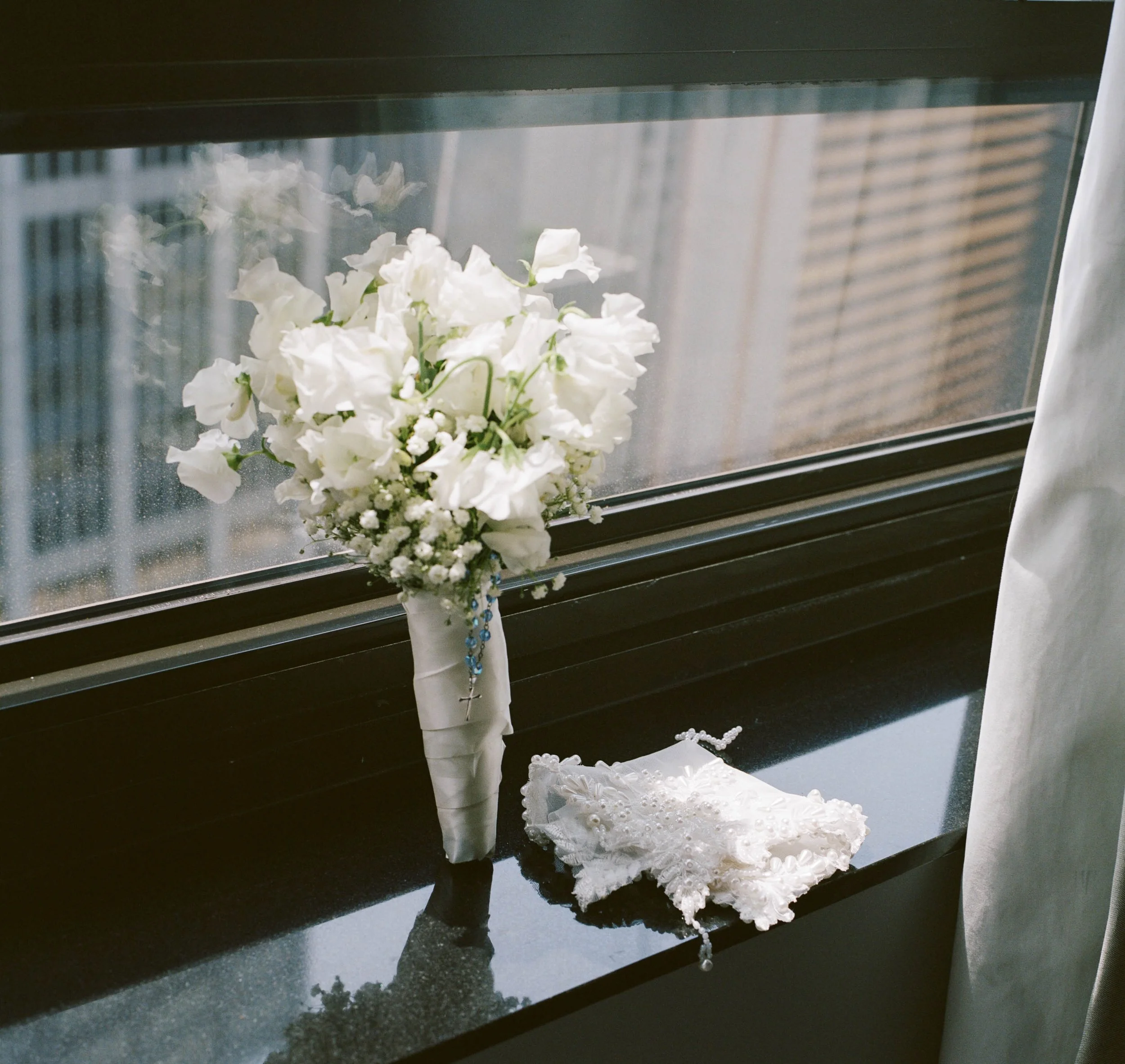 A white bouquet of flowers and a white lace handkerchief or cloth on a black windowsill, with a city view and rain outside the window.