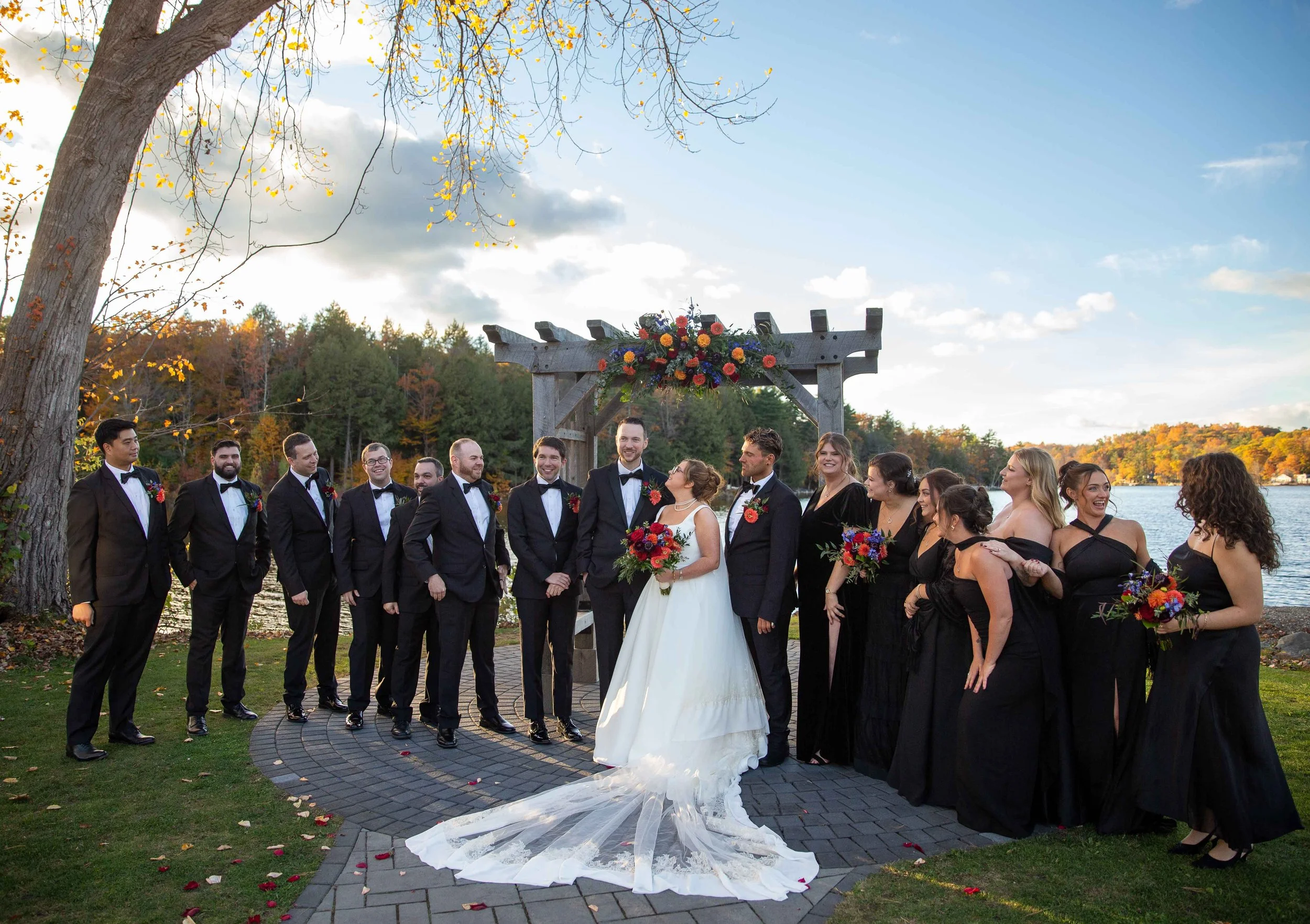 A wedding party posing outdoors by a lake, with a wooden arch decorated with flowers in the background. The bride is in a white wedding dress holding a bouquet, surrounded by groomsmen in black suits and bridesmaids in black dresses holding bouquets.