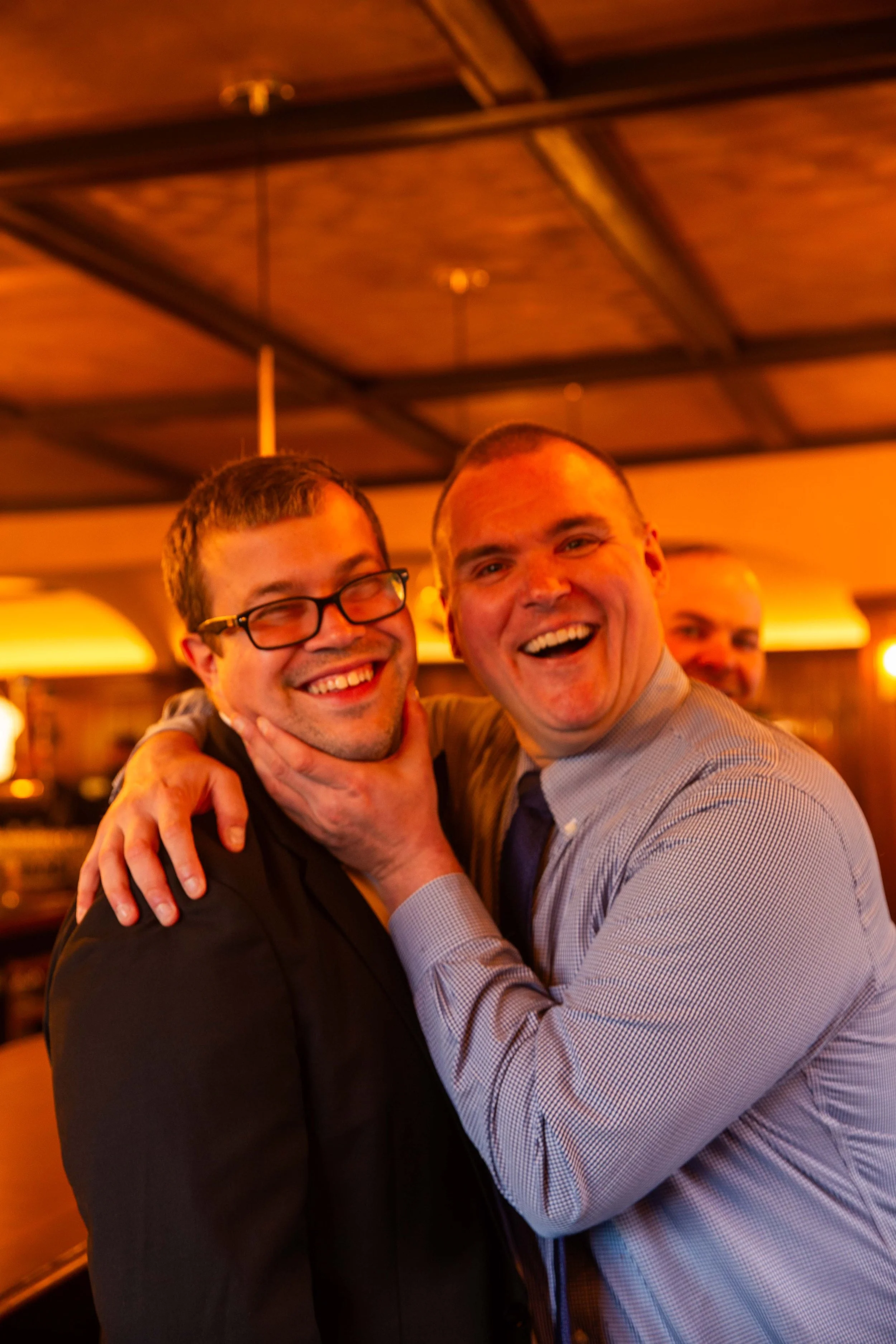 Two men smiling and hugging in a warm-lit indoor setting, one wearing glasses and the other without, with a third man visible in the background.