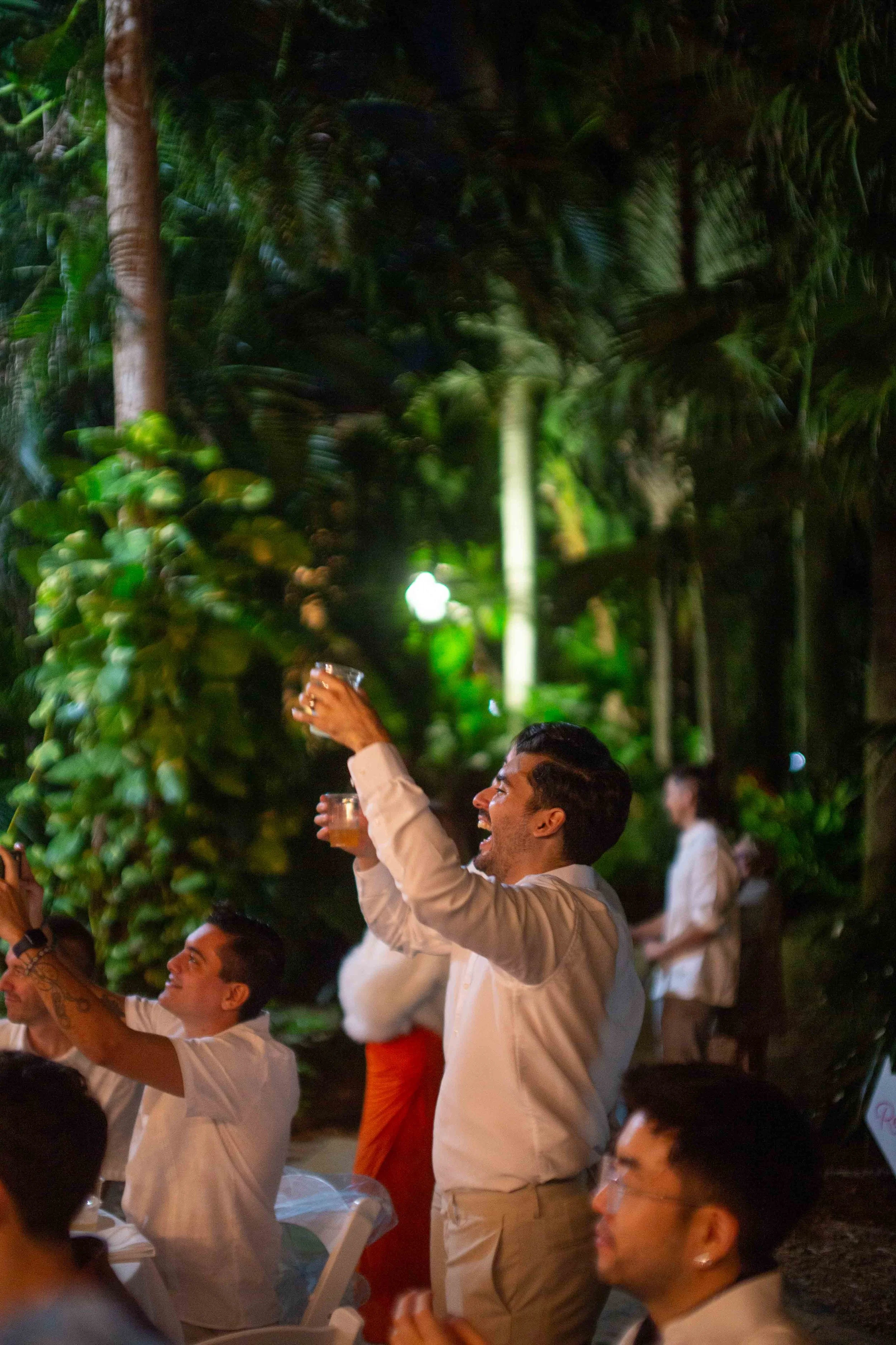 People raising glasses at an outdoor celebration in a lush, tropical setting during nighttime.