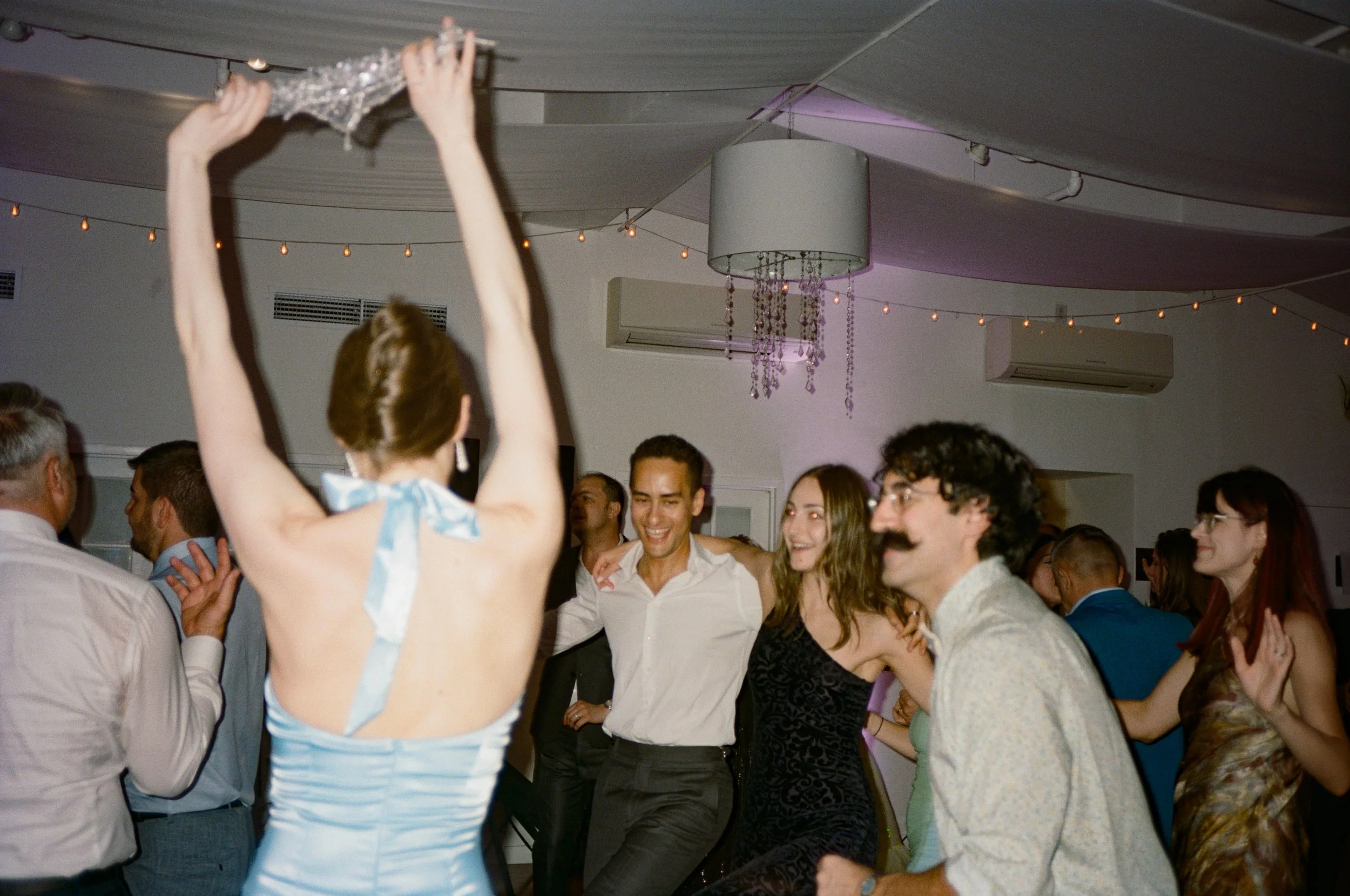 People dancing and enjoying a party in an indoor venue with string lights and a chandelier.