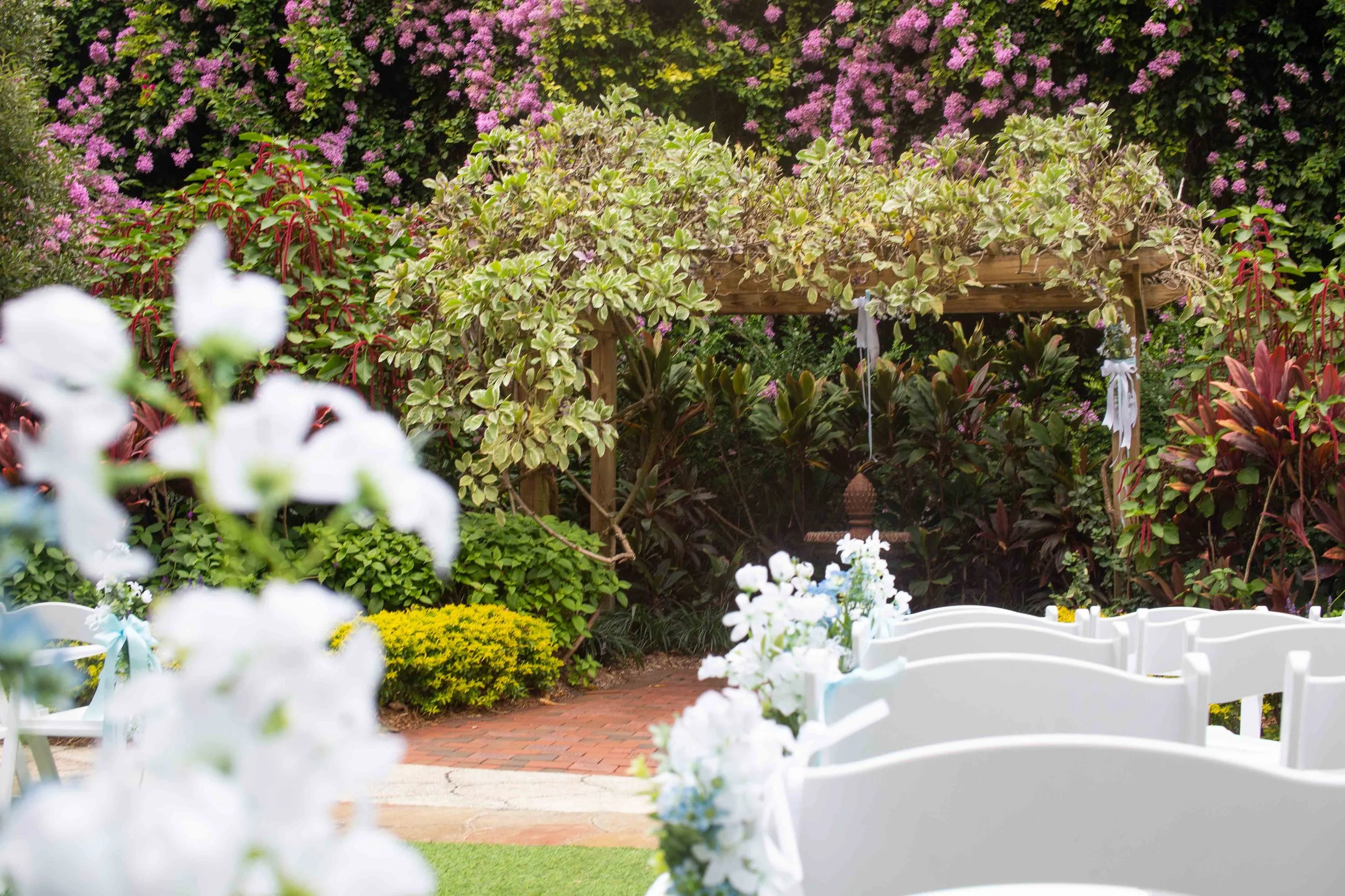 Outdoor wedding setup with white chairs decorated with flowers, a wooden arbor with plants, and a garden backdrop with blooming flowers.