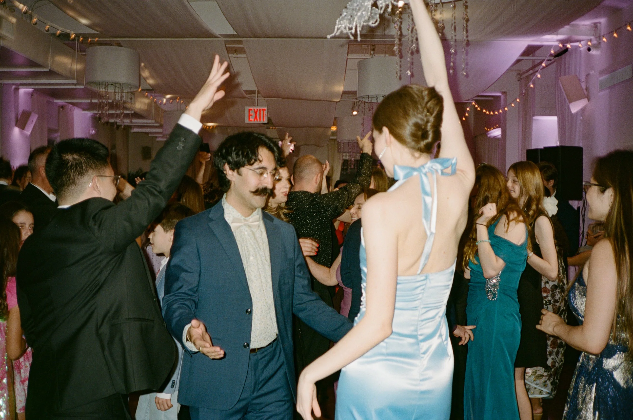 People dancing at a formal event or party in a decorated hall with pink lighting, chandeliers, and string lights.