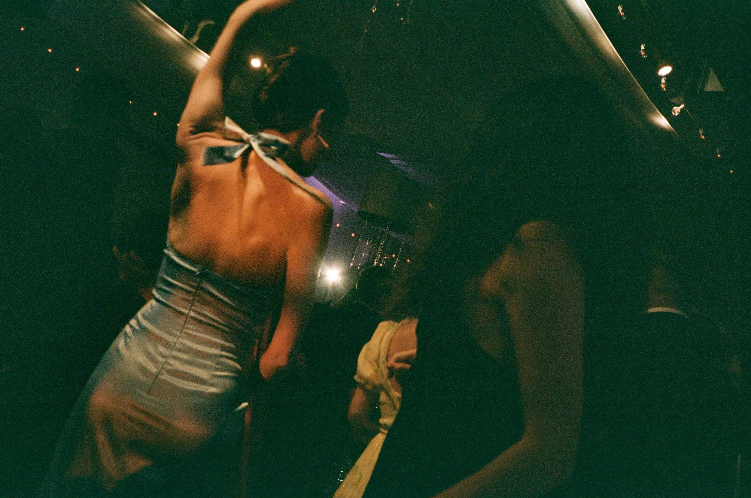 A woman in a black top and yellow shorts interacting with a woman in a silver dress inside a dark, possibly nightclub setting.
