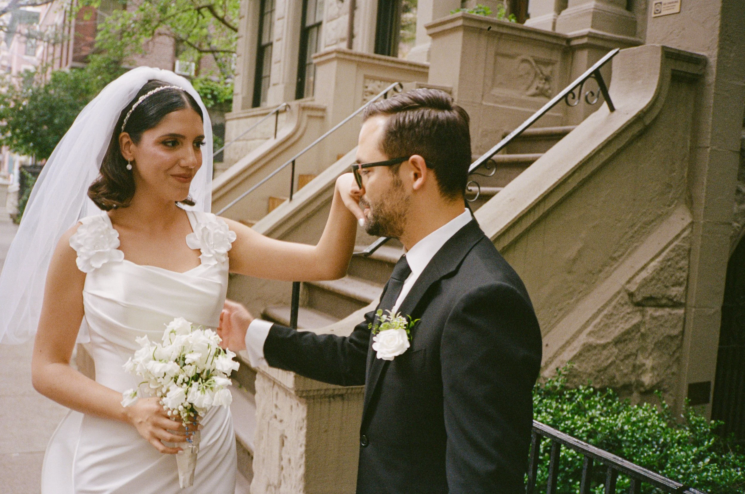 A bride and groom on a city street, the bride with dark hair wearing a white dress and veil, holding a bouquet of white flowers, gently touching the groom's face. The groom with glasses, beard, in a black suit, white shirt, and boutonniere, is touchi