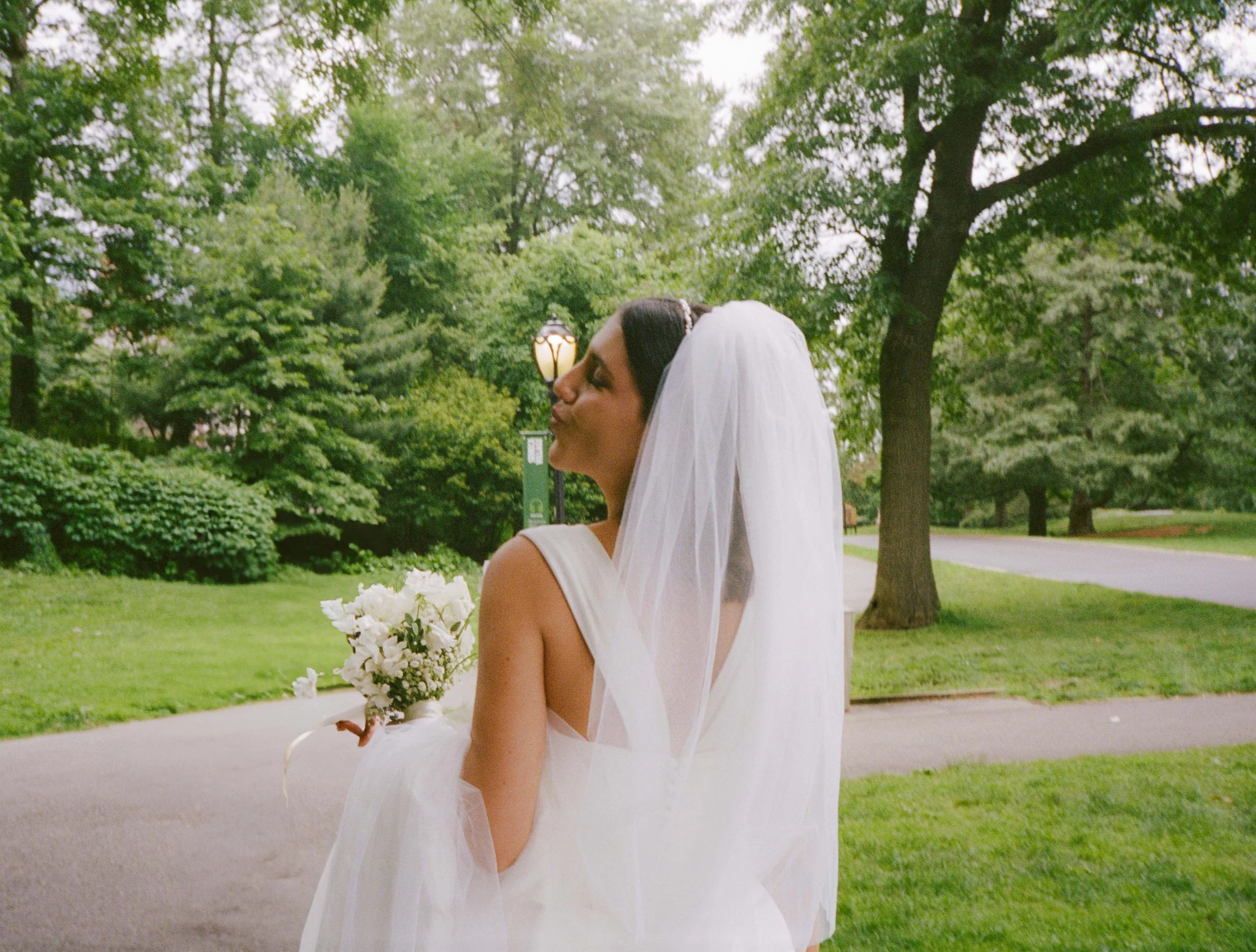 Bride in a white wedding dress with a veil holding a bouquet of white flowers in a park with green trees and a lamppost