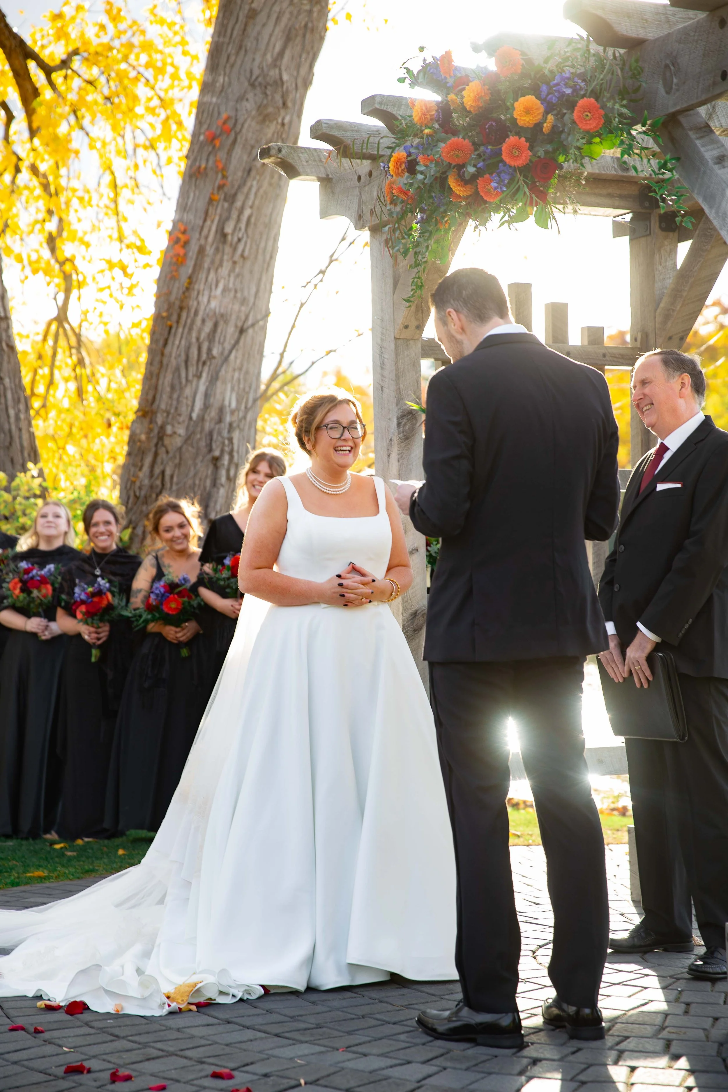 Bride and groom exchanging vows during outdoor wedding ceremony under a wooden arch decorated with a colorful floral arrangement, with bridesmaids and guests watching.