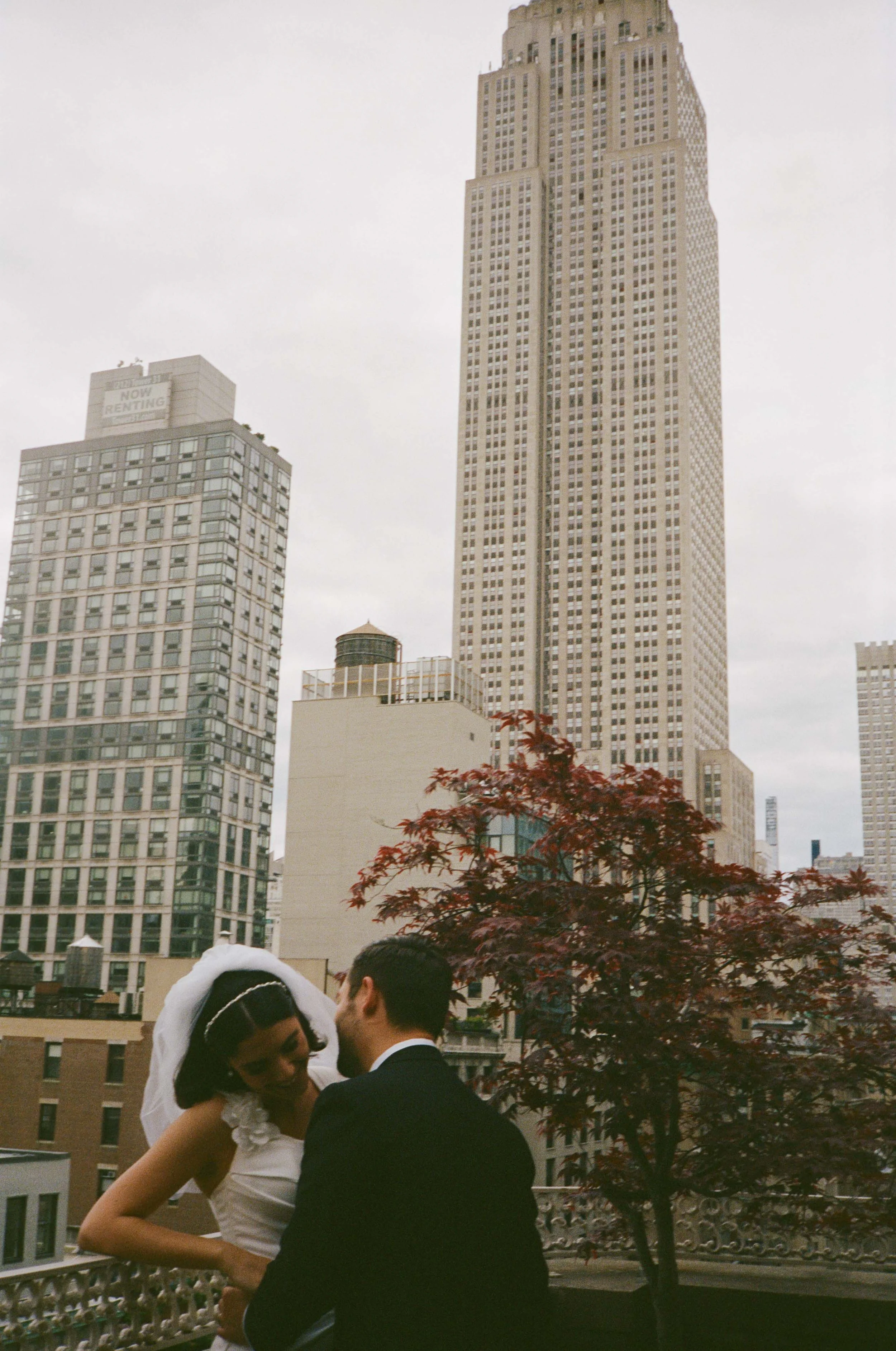 A bride and groom share a kiss or embrace on a balcony with tall city buildings and a red-leaved tree in the background.