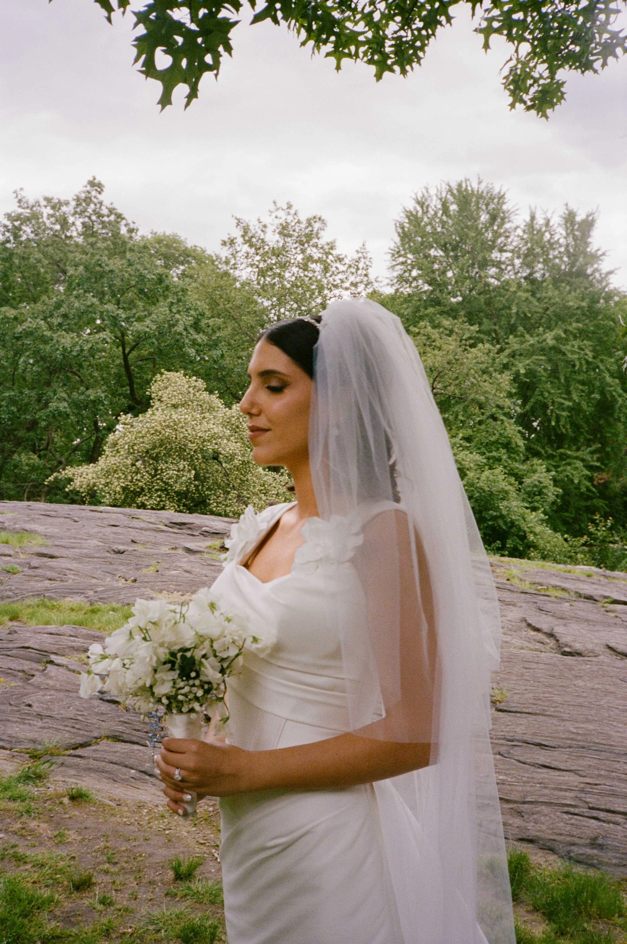 A bride in a white wedding dress and veil holding a bouquet of white flowers outdoors with greenery and rocks in the background.