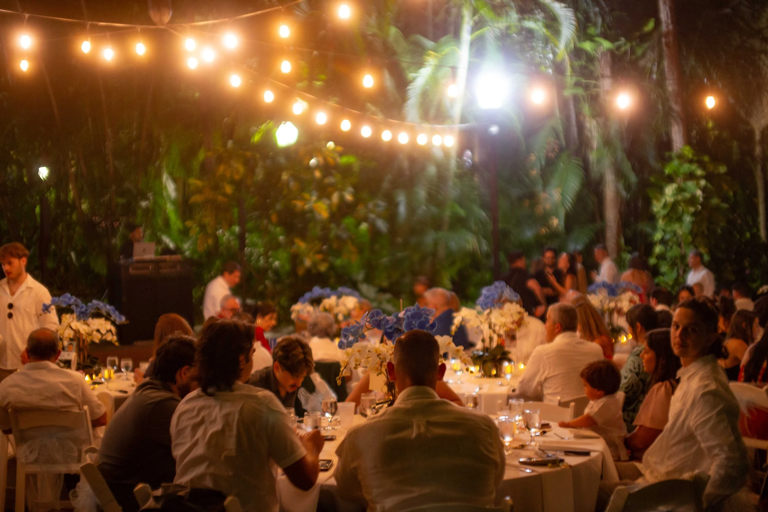 People dining at a nighttime outdoor event under string lights in a lush garden setting.