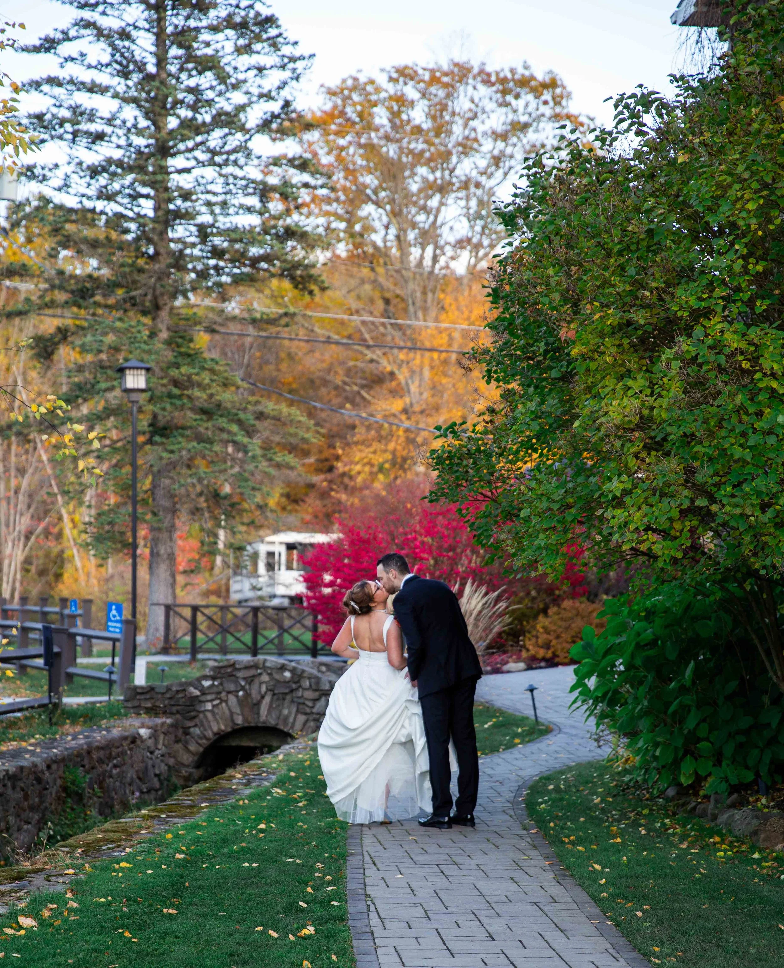 A bride and groom sharing a kiss on a stone pathway in a park during autumn, surrounded by colorful fall foliage.