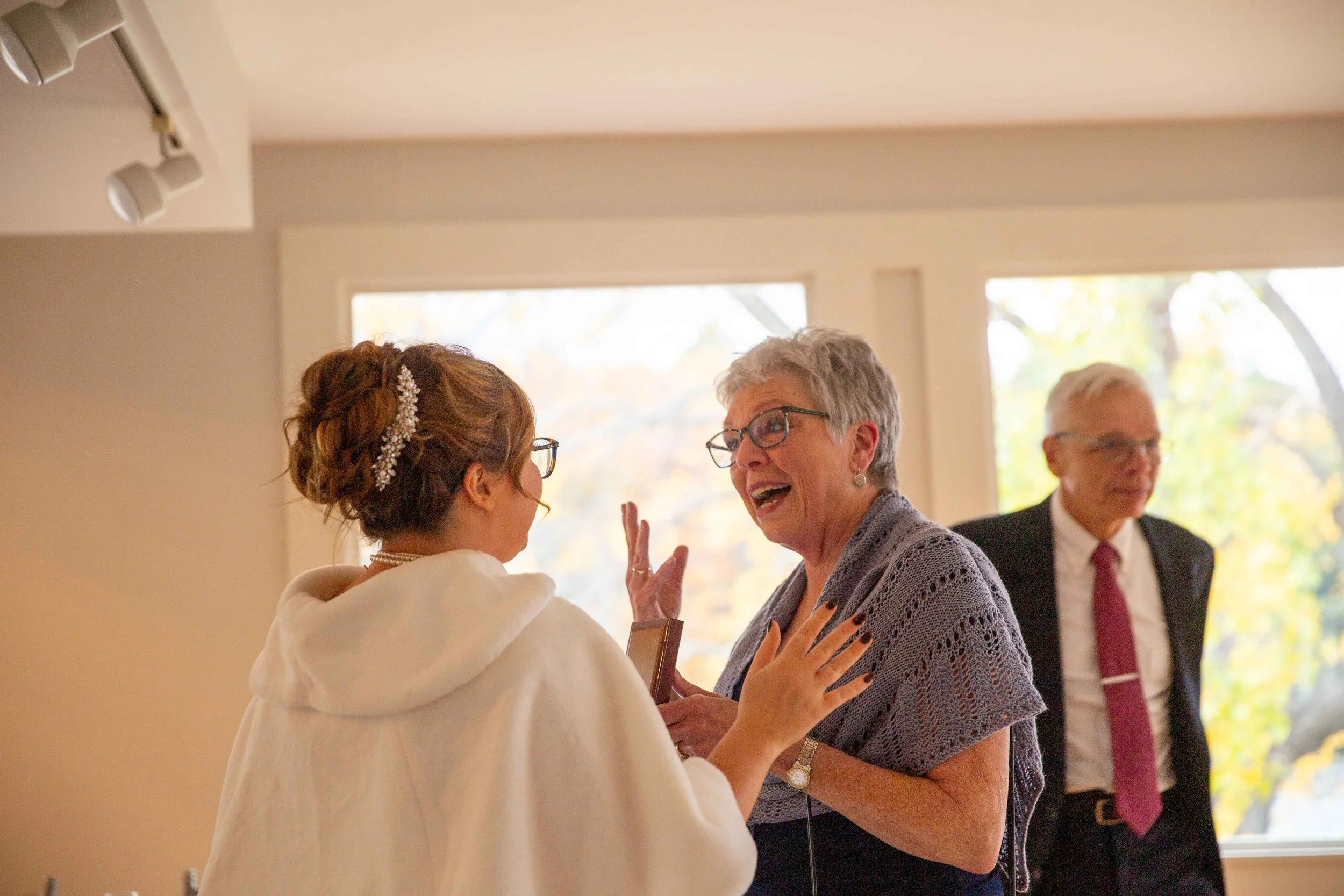 A wedding scene with a young bride in white dress and older woman in grey shawl sharing a joyful moment, while a man in suit looks on in background.