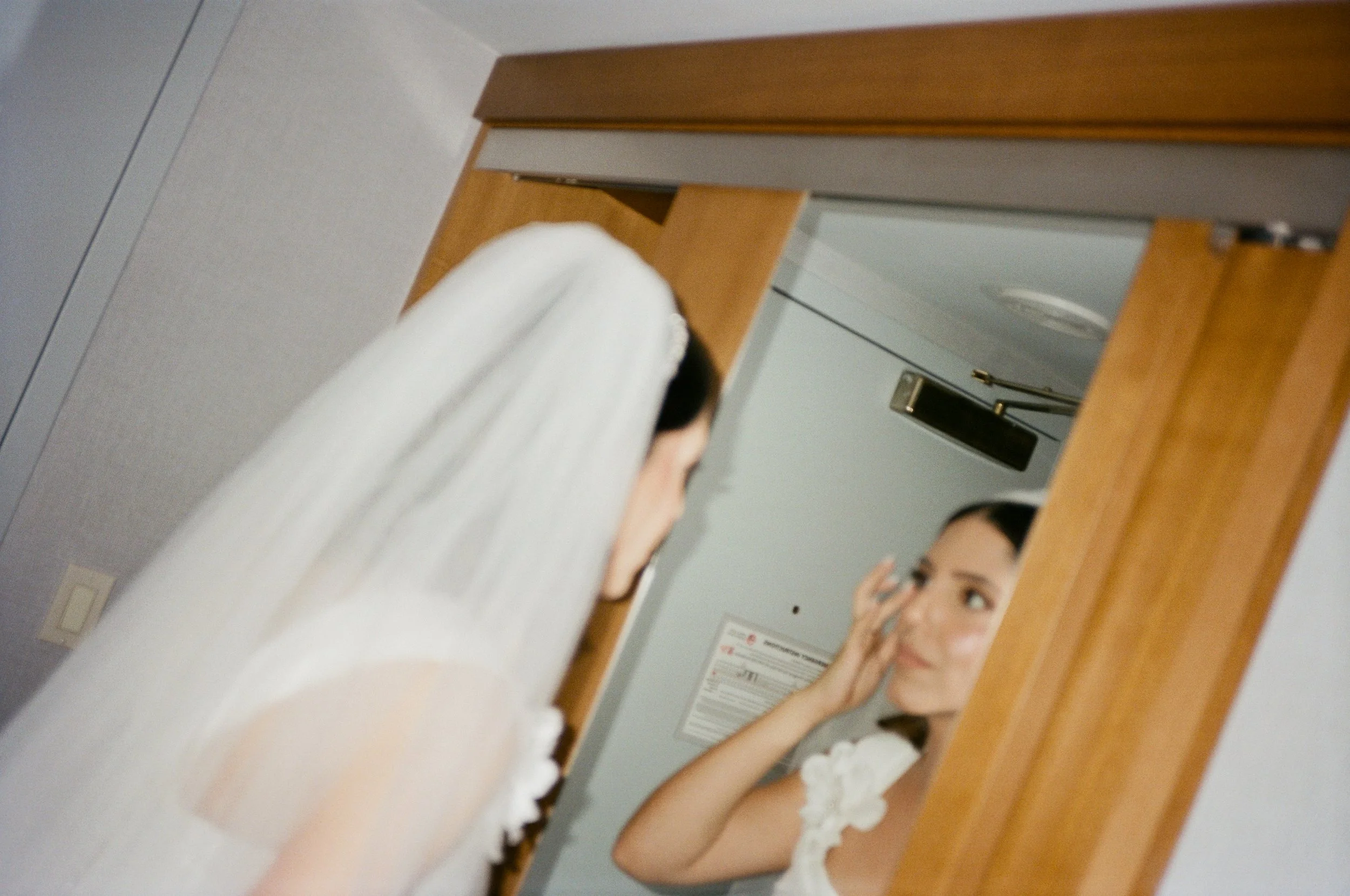 A woman in a white wedding dress and veil is looking at herself in a mirror, touching her face.