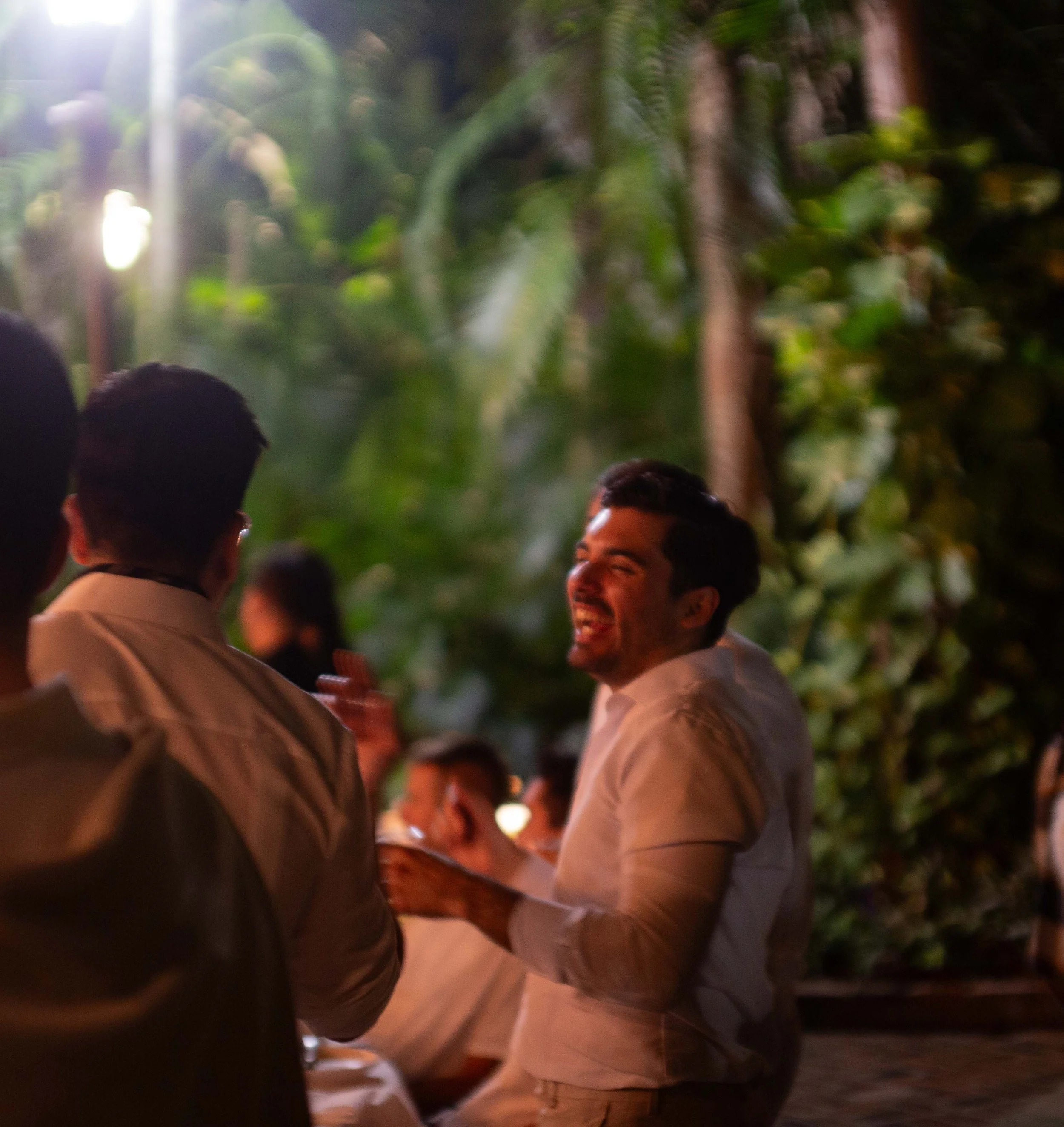 People socializing outdoors at night, surrounded by lush greenery, with one person laughing.