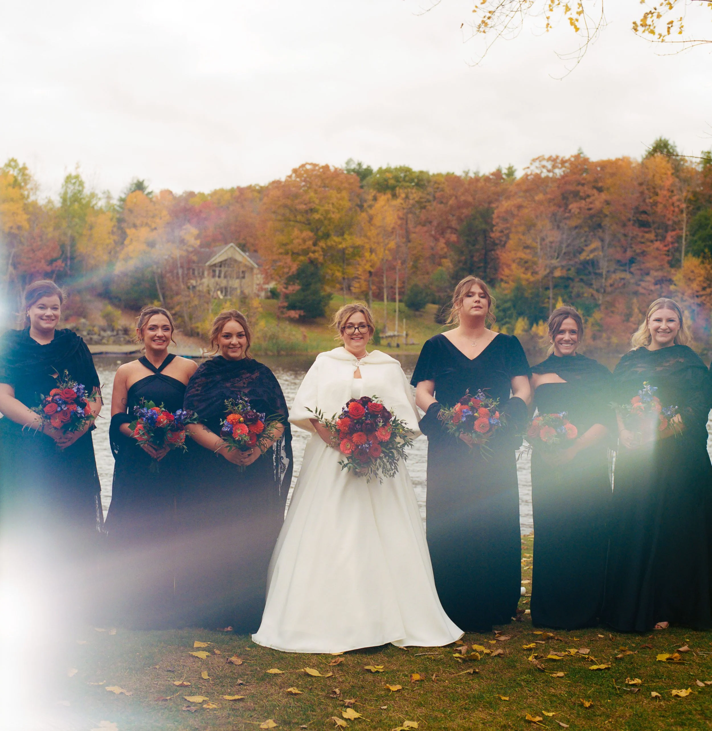 Group of women in formal black dresses, with one woman in a white wedding gown, standing outdoors near a lake with fall foliage in the background, holding bouquets of flowers.