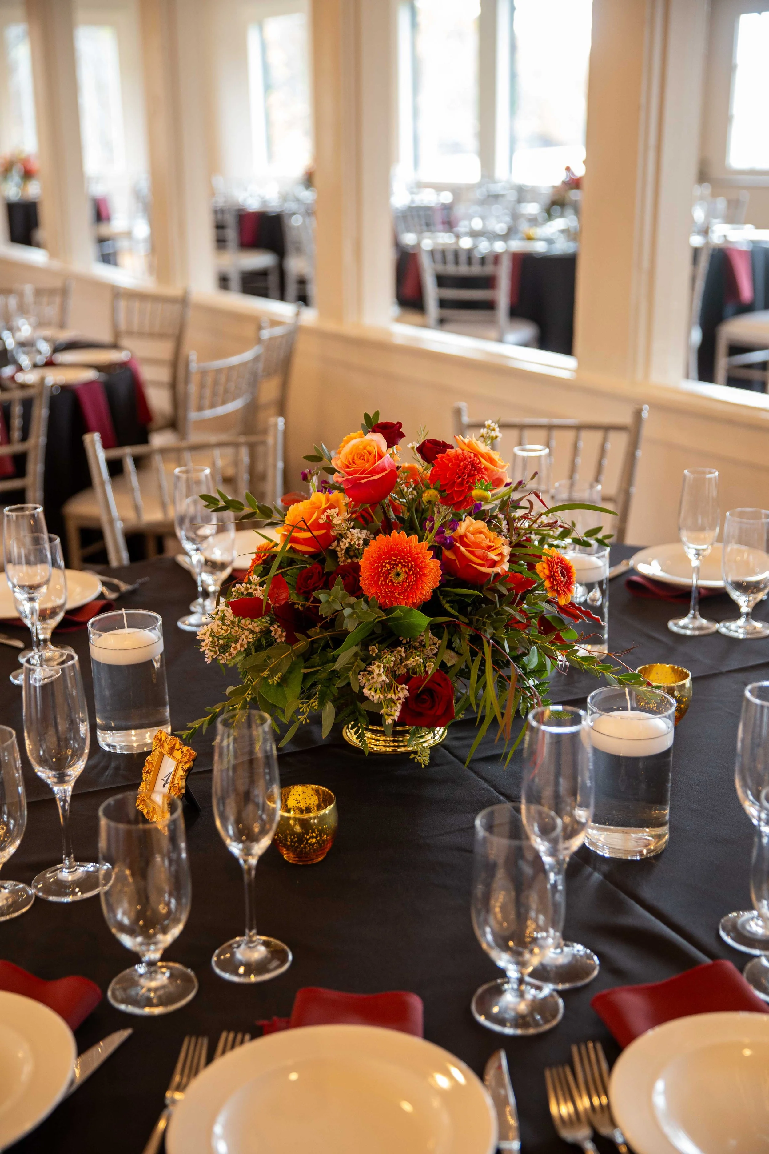 Elegant restaurant banquet table with a large colorful floral centerpiece, surrounded by water glasses, candles, and place settings with white plates, silverware, and red cloth napkins, in front of large windows letting in natural light.