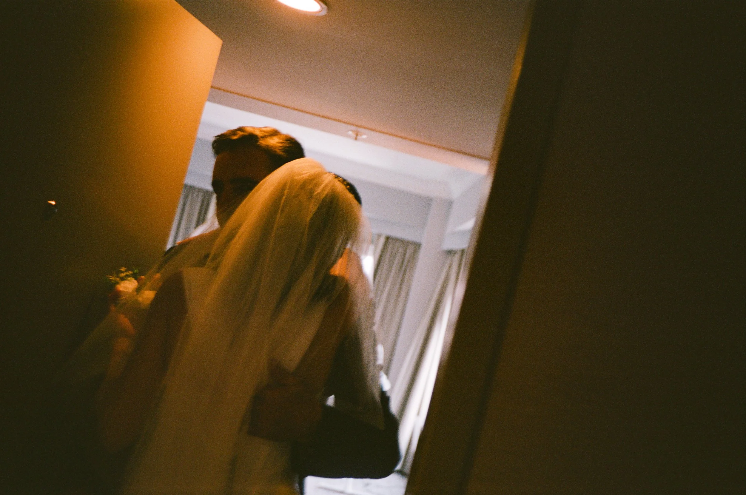 A woman in a wedding dress and veil, and a man in a suit, are seen through a reflection on a mirror, likely during a wedding or formal event.