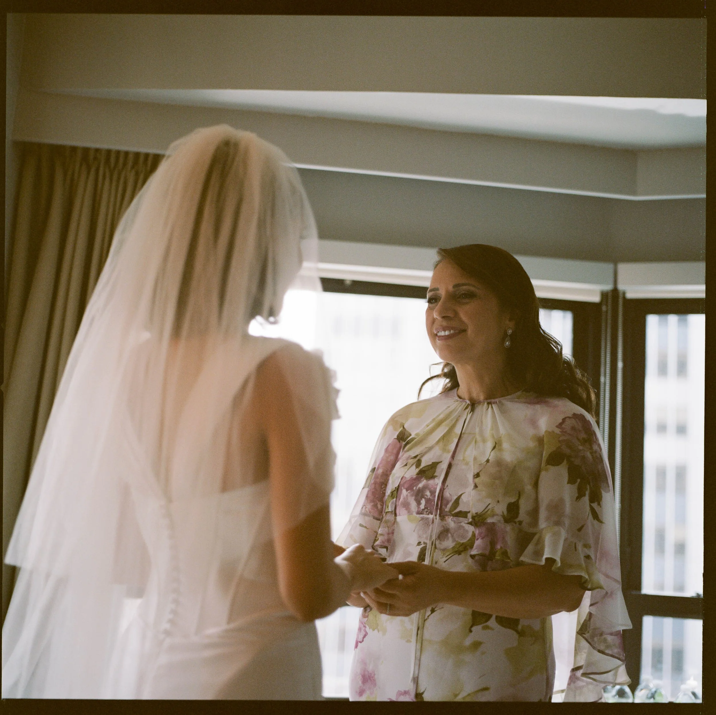A bride in a wedding dress with a veil holding hands with a woman in a floral dress, possibly her mother, in a room with large windows.
