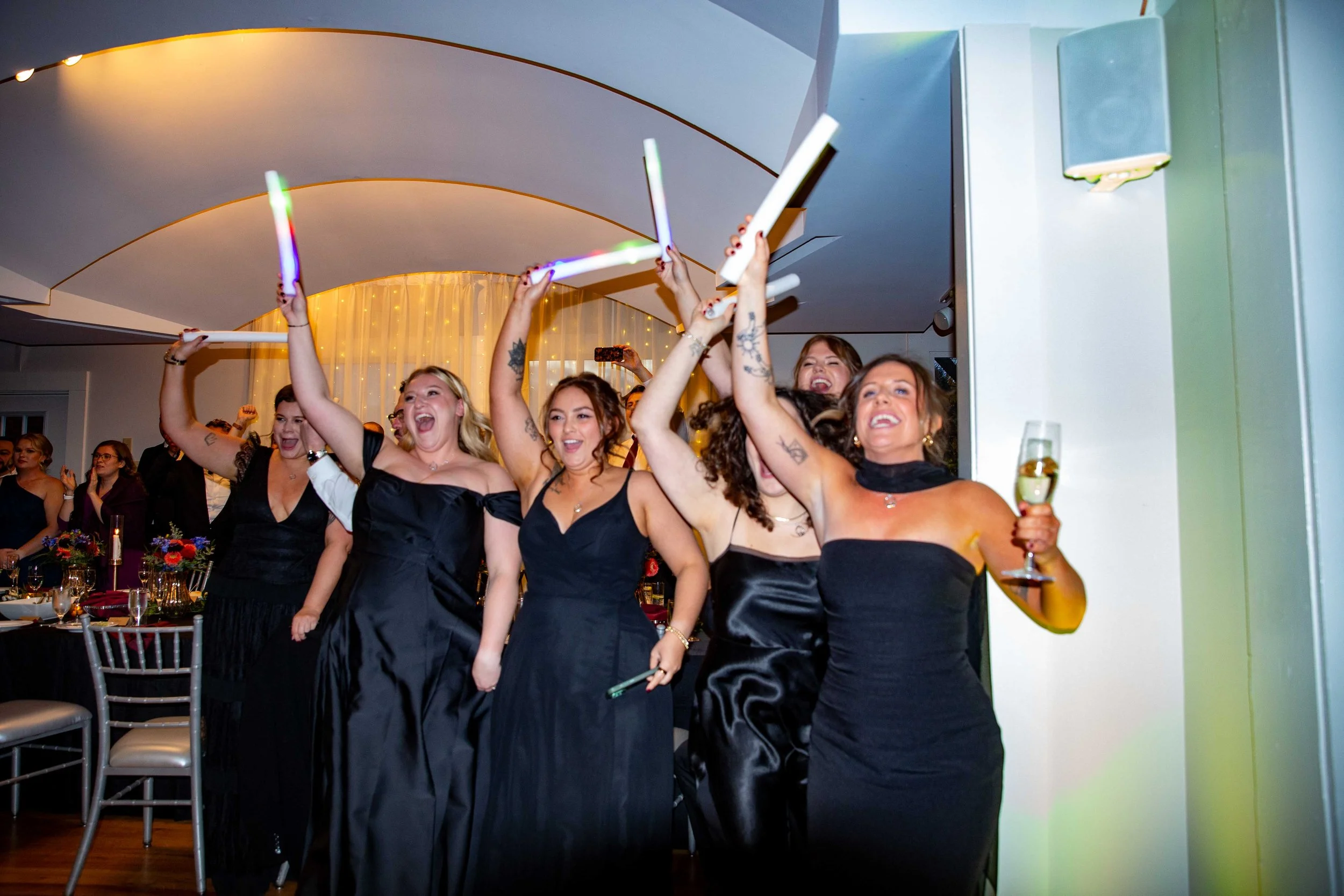 Group of women celebrating at an event, holding glow sticks and a drink, dressed in black formal dresses.
