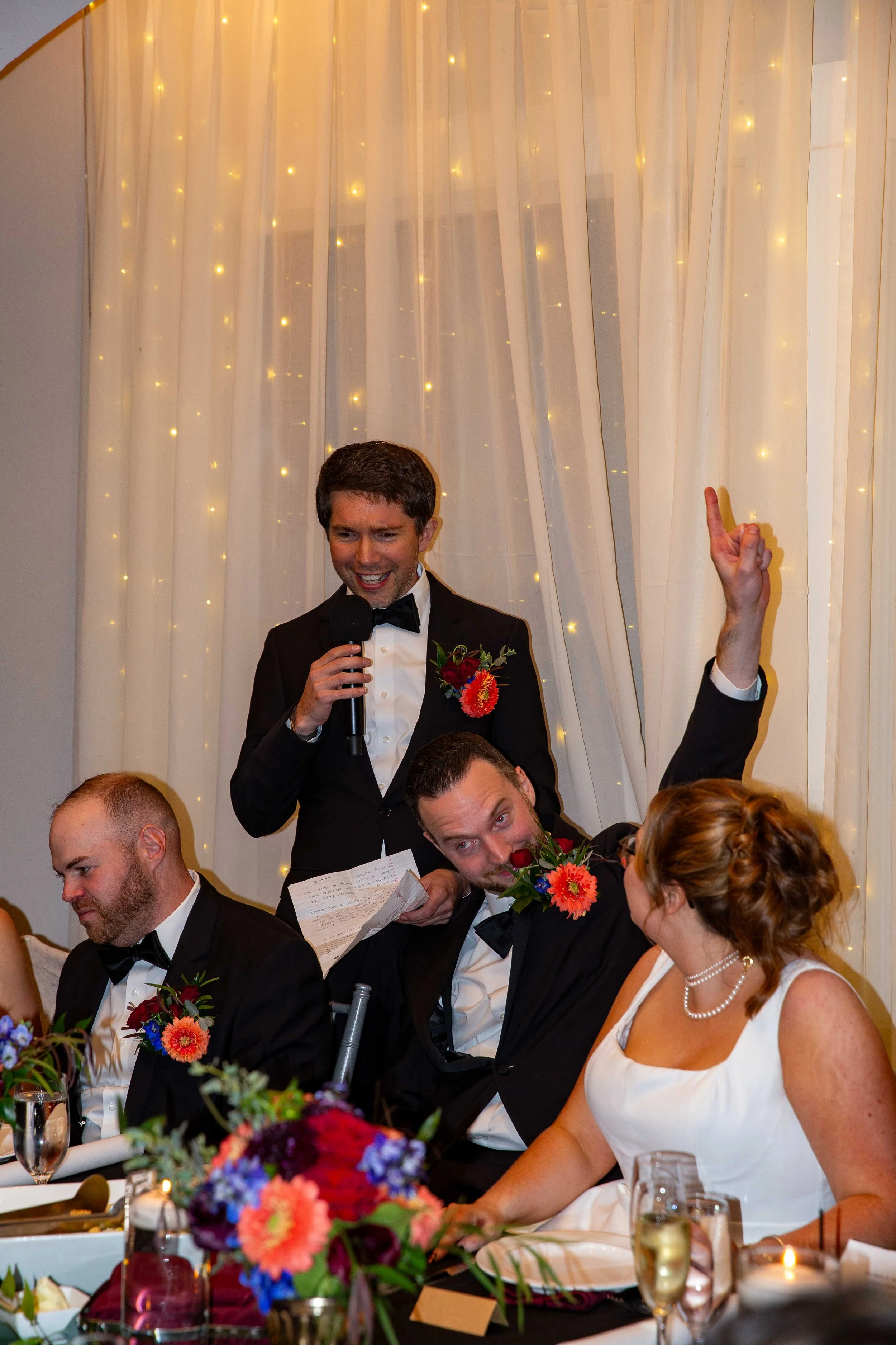 A man in a tuxedo giving a speech at a wedding reception, with others seated at the table listening. The setting is decorated with fairy lights and flowers.