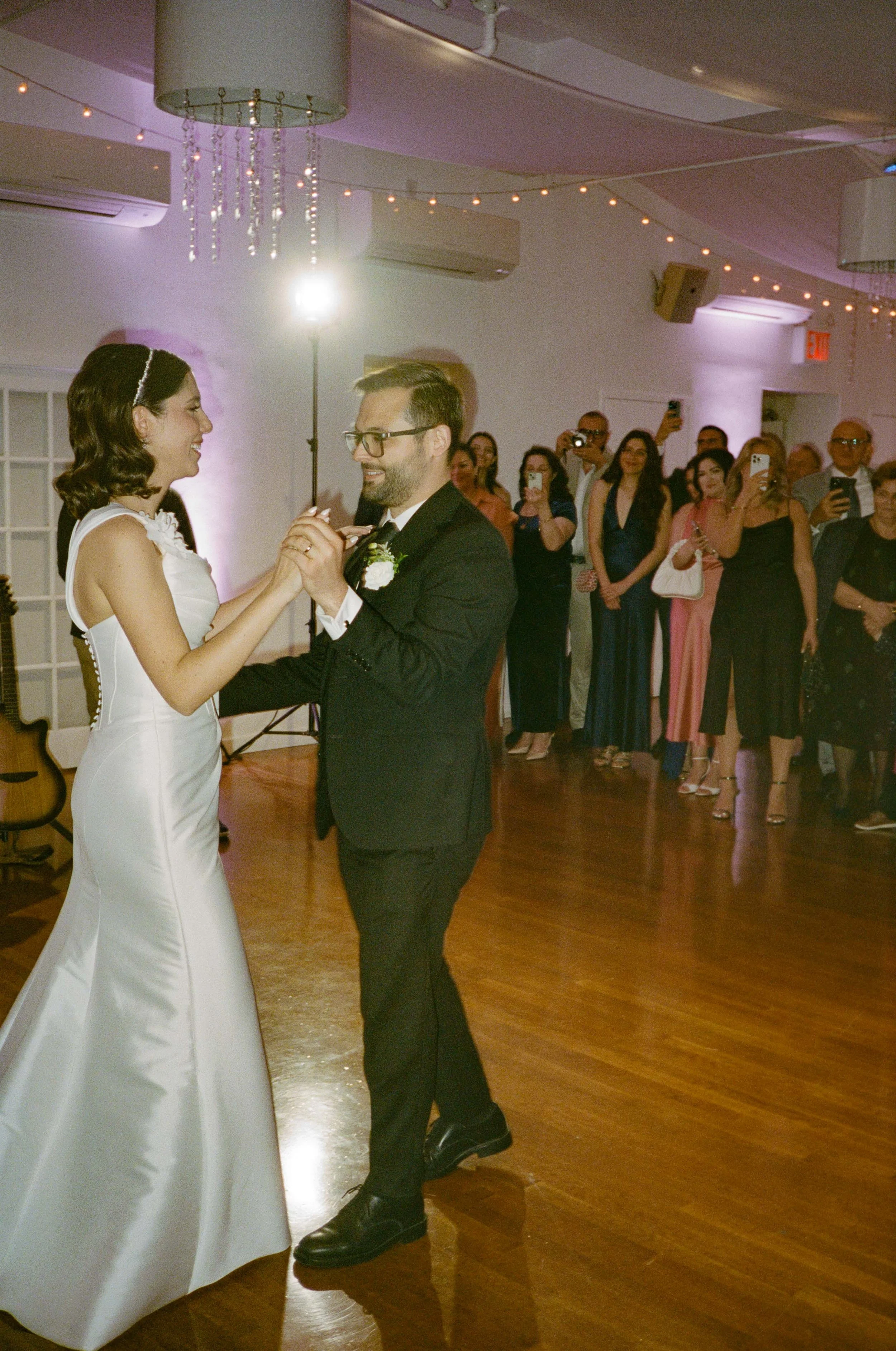 A bride and groom sharing their first dance during a wedding reception, with guests watching and taking photos in the background.