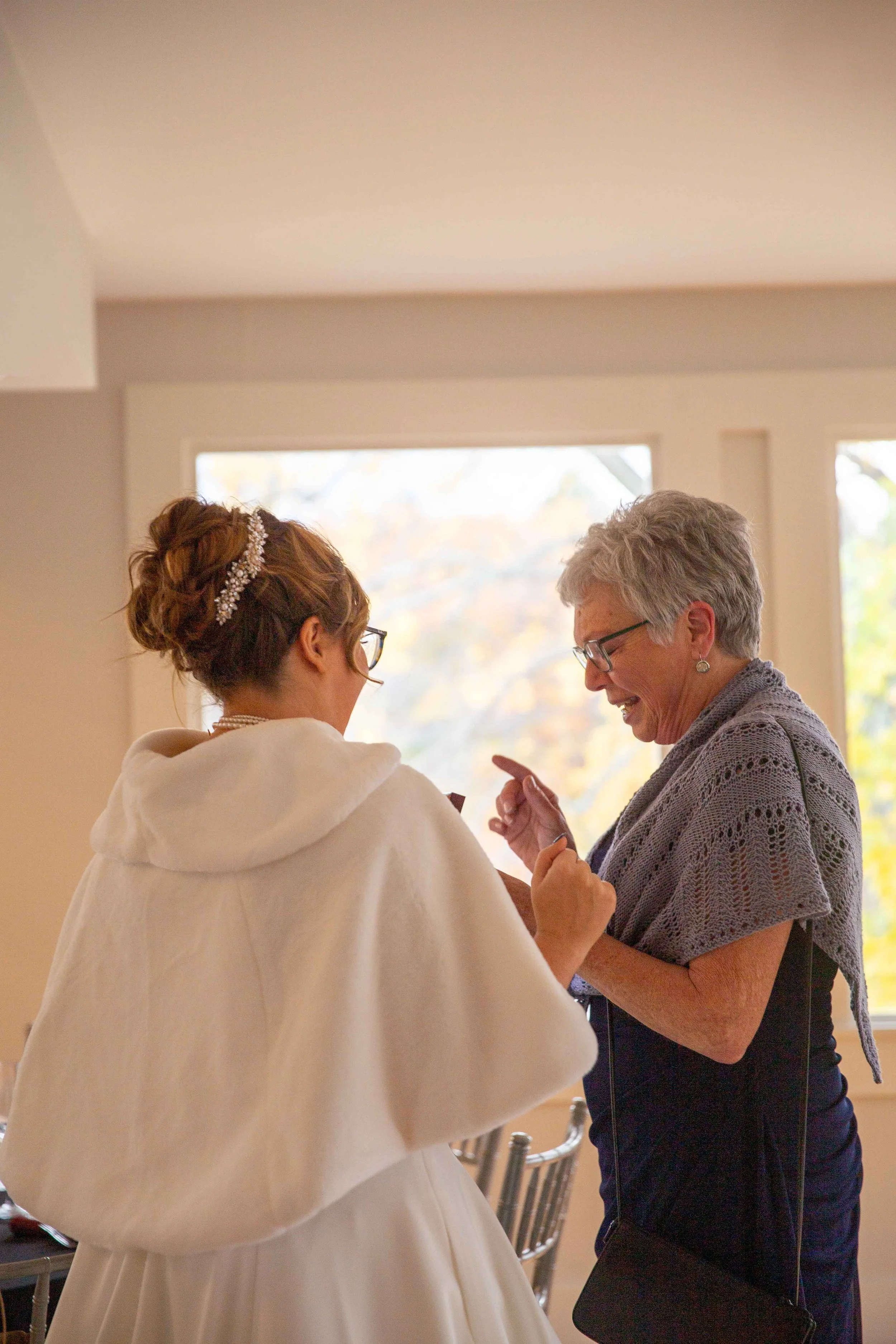 A bride with an updo hairstyle and glasses smiles and interacts with an older woman with short gray hair and glasses inside a room with large windows showing autumn foliage outside.