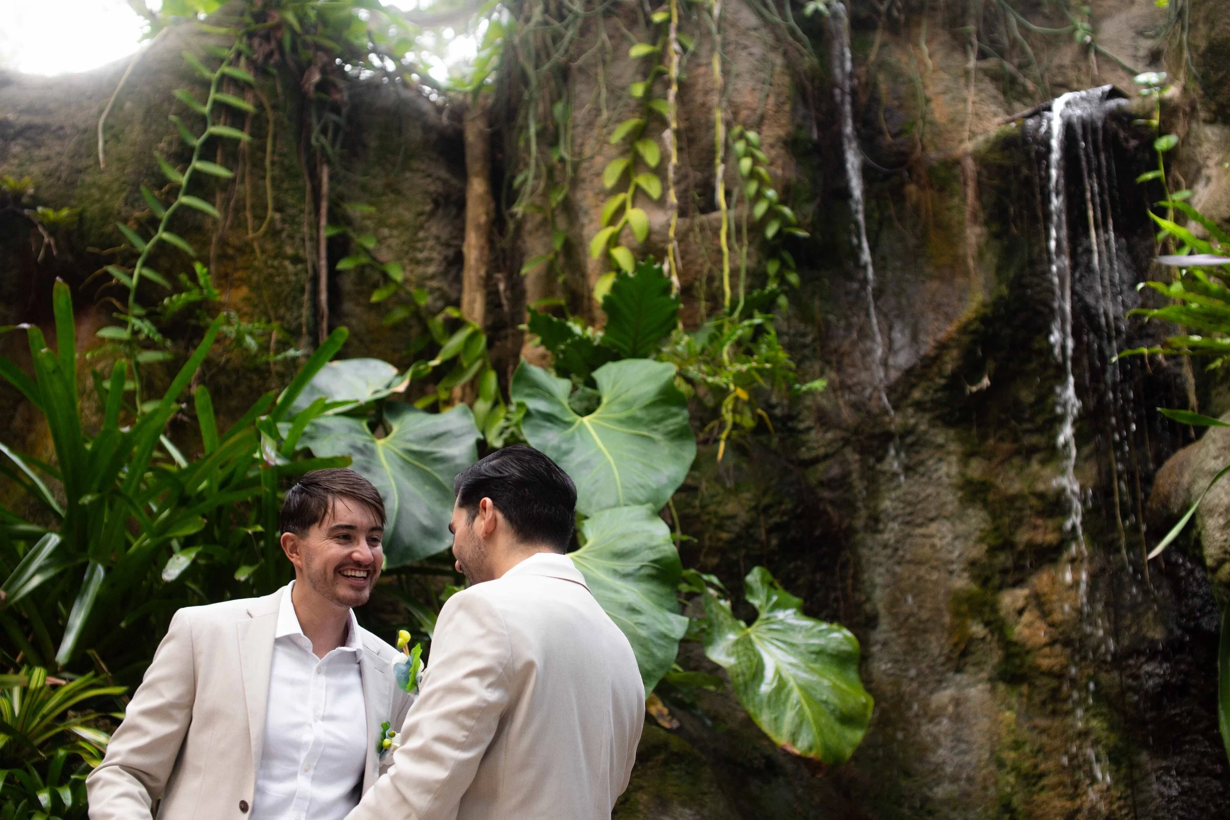 Two men in wedding attire smiling and facing each other in front of a lush green garden with large leaves and a small waterfall.