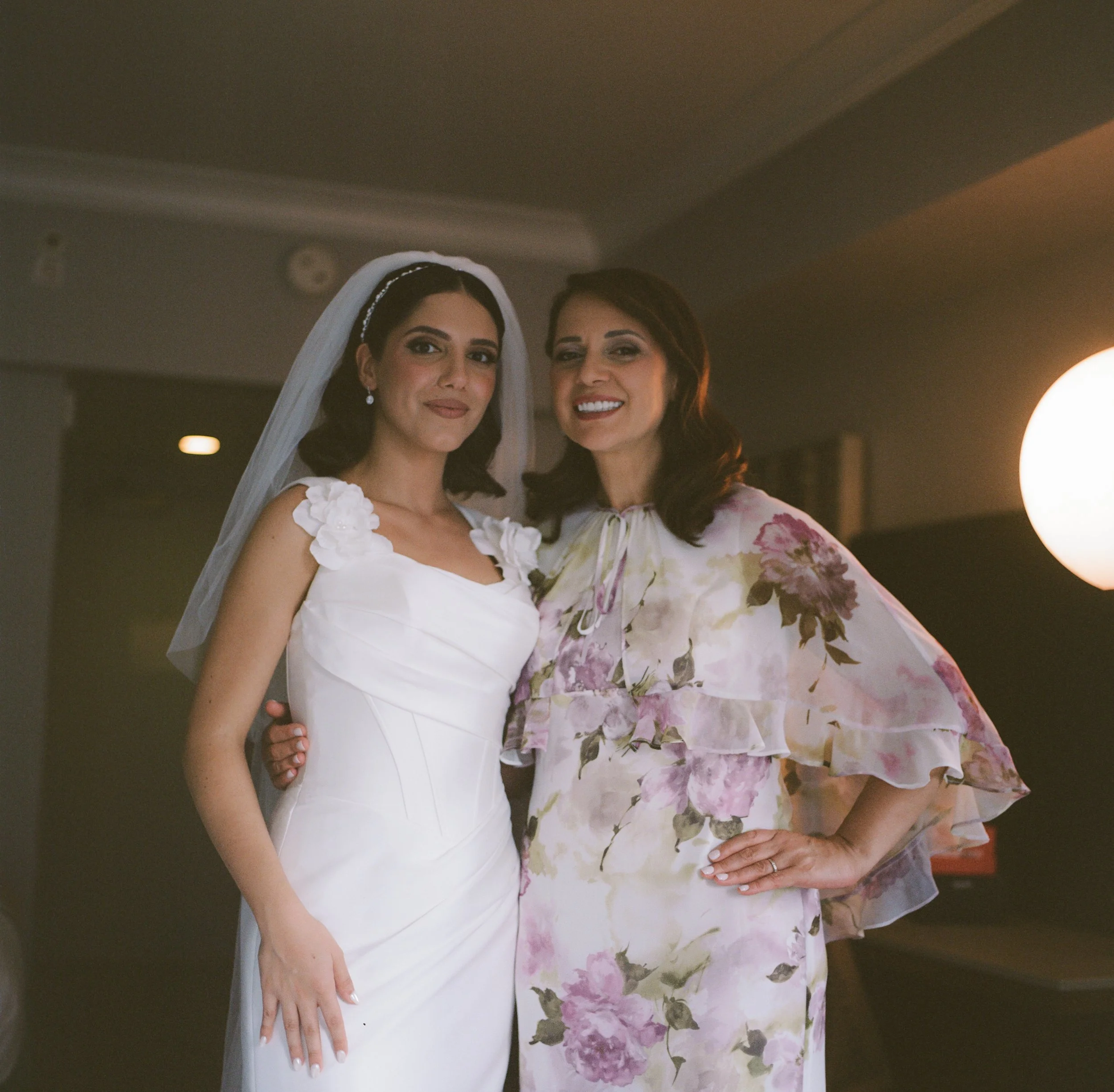 A bride in a white wedding dress with floral details on the shoulders, wearing a veil, standing next to a woman in a floral dress, both smiling at the camera indoors.
