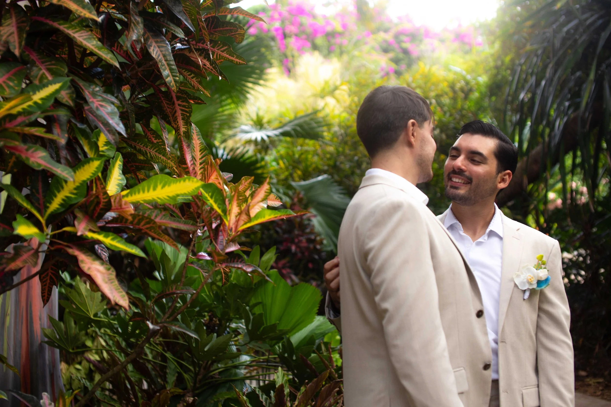 Two men in cream-colored suits sharing a special moment in a lush tropical garden, smiling and holding each other close.