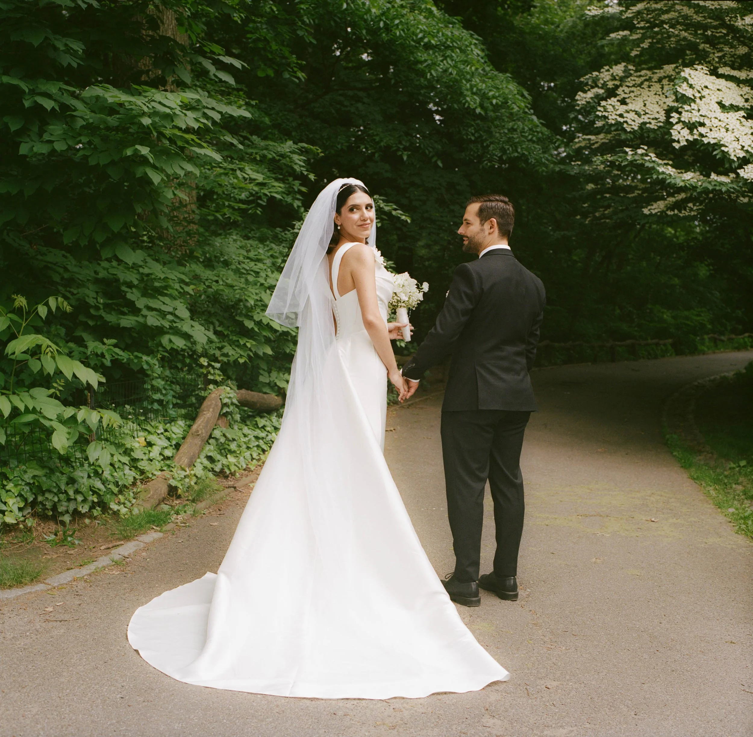 Bride and groom holding hands on a forest path, the bride in a white wedding gown and veil, holding a bouquet, the groom in a black suit, surrounded by green trees.