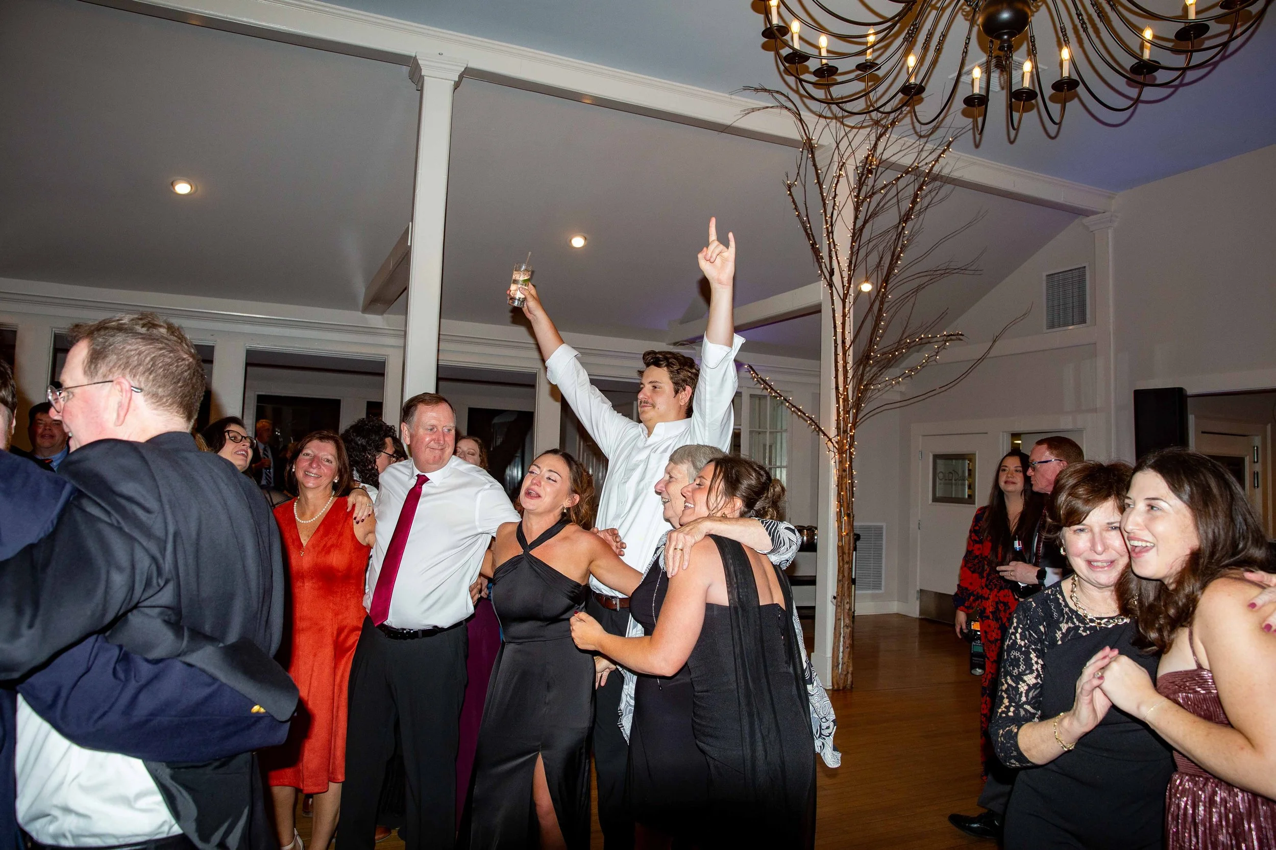 A group of people dancing and celebrating at a party or wedding reception, with one man holding a drink and raising his hand.