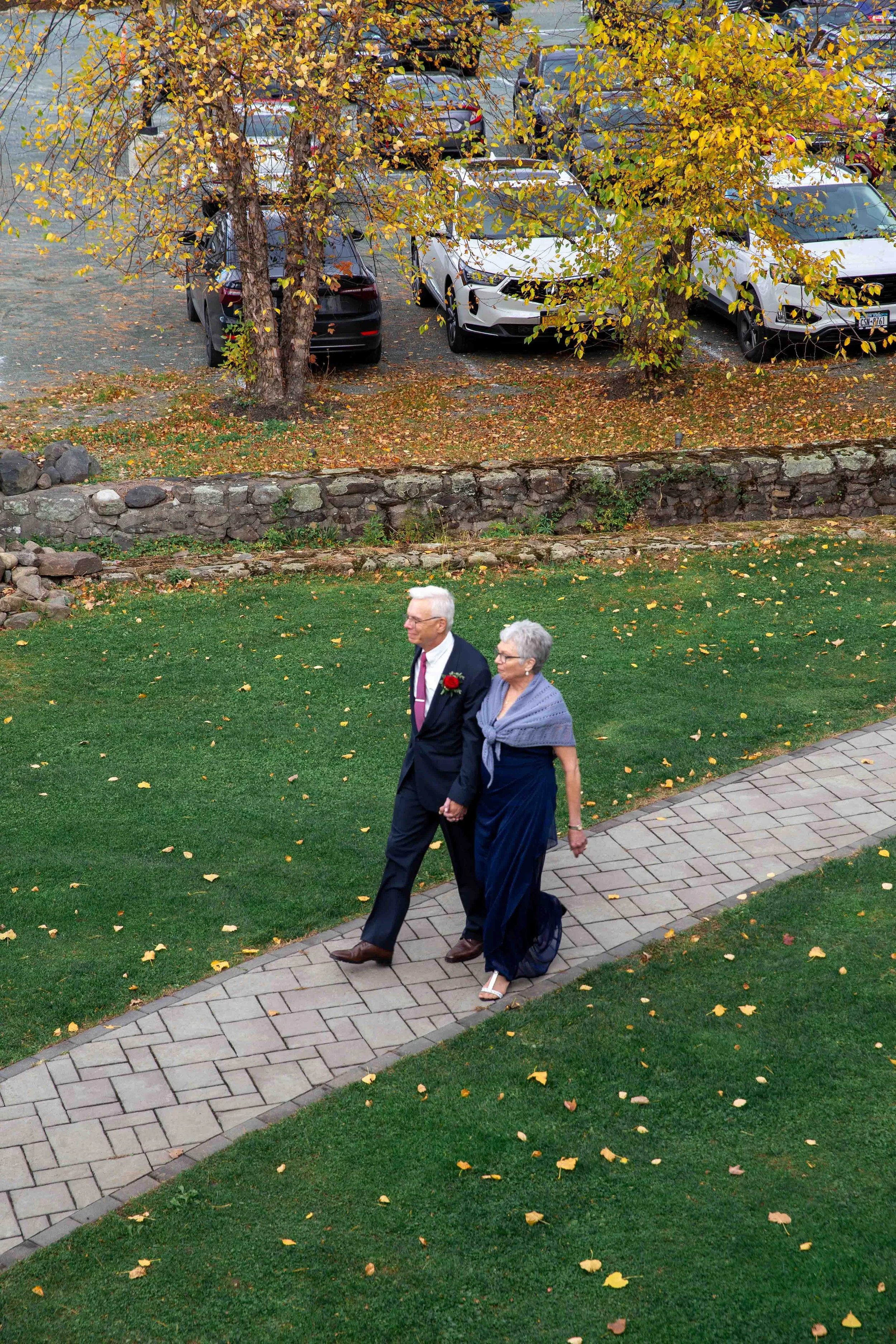 An elderly couple walking hand in hand on a brick pathway, with the man dressed in a suit and the woman in a blue dress and shawl, surrounded by fallen autumn leaves and green grass, with parked cars and trees with yellow and brown leaves in the back