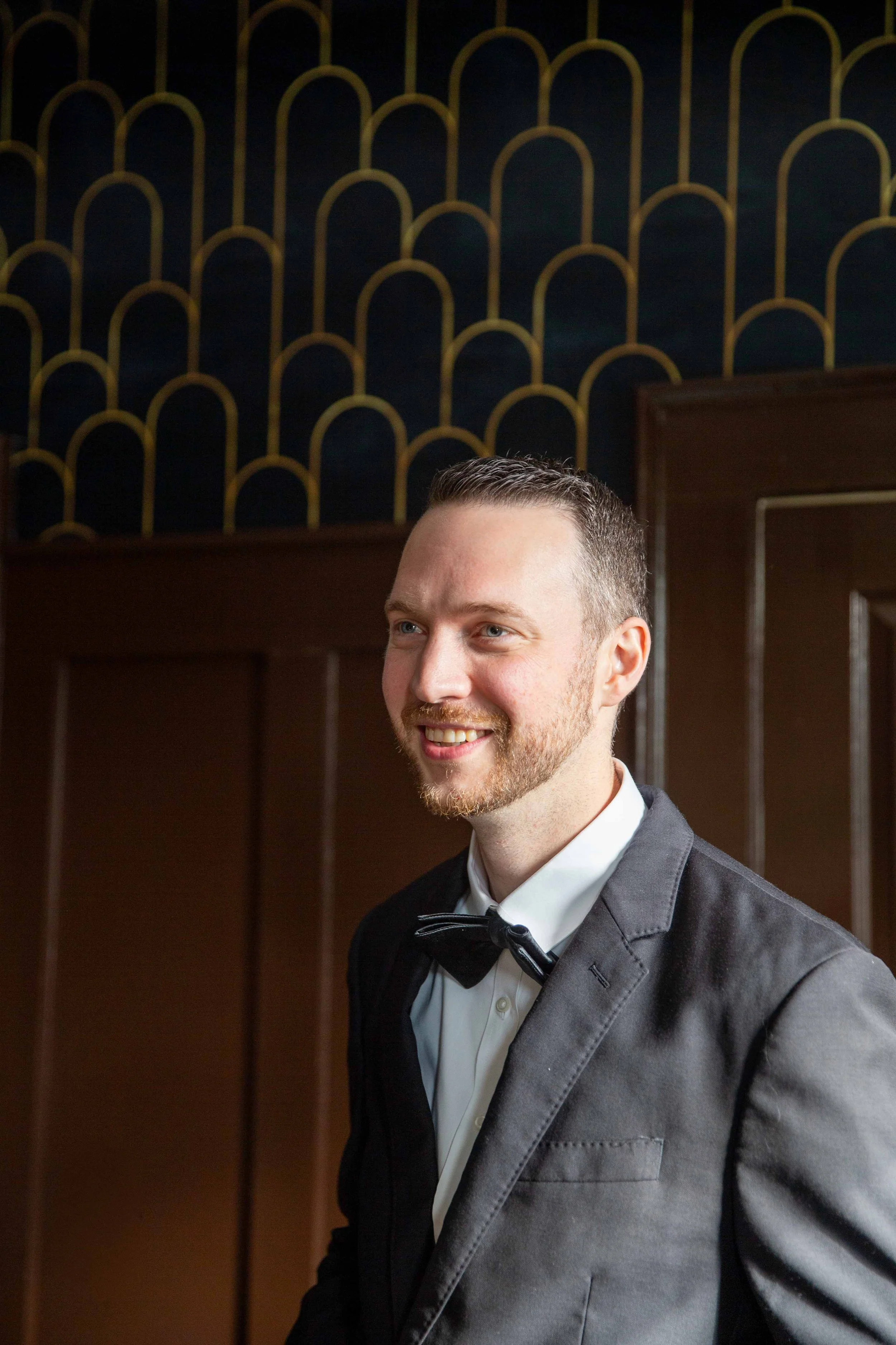 A man in a tuxedo with a bow tie smiling indoors, standing in front of a dark background with gold arch patterns.