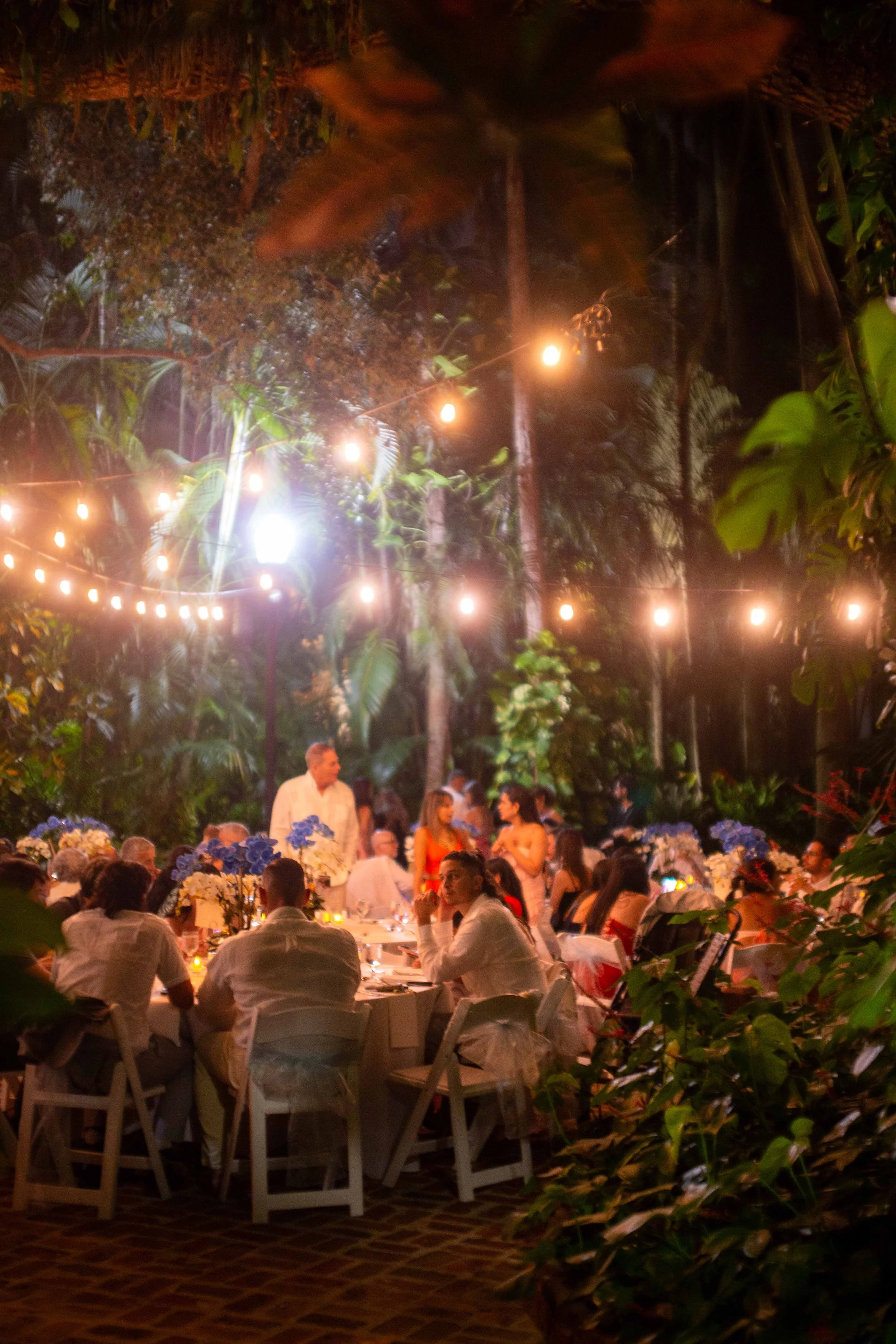 People gathered at an outdoor evening event, sitting at tables under string lights in a lush, tropical garden setting.