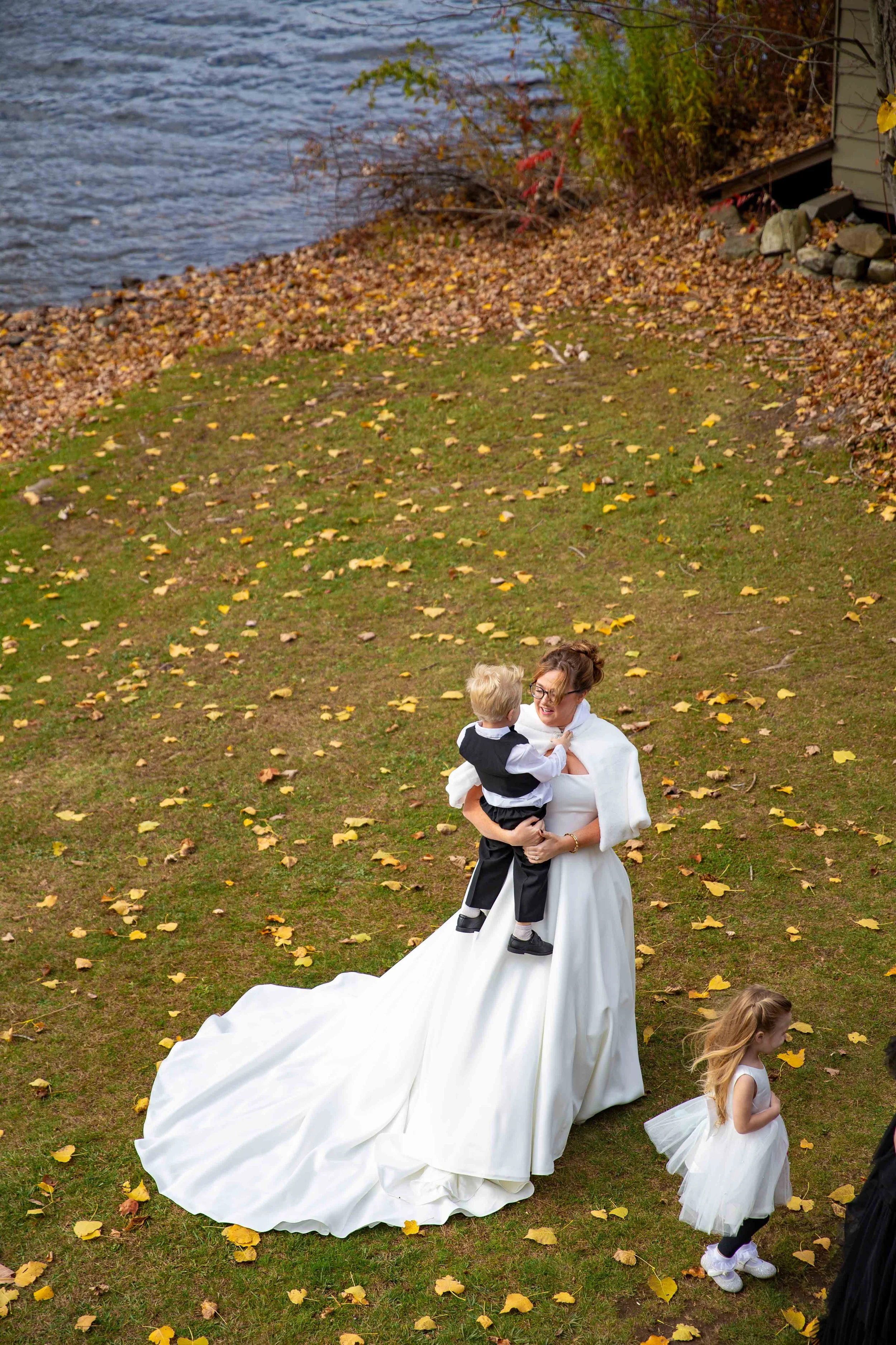 A woman in a white wedding dress holding a young boy in formal attire outdoors near a body of water with fallen yellow leaves on the ground.