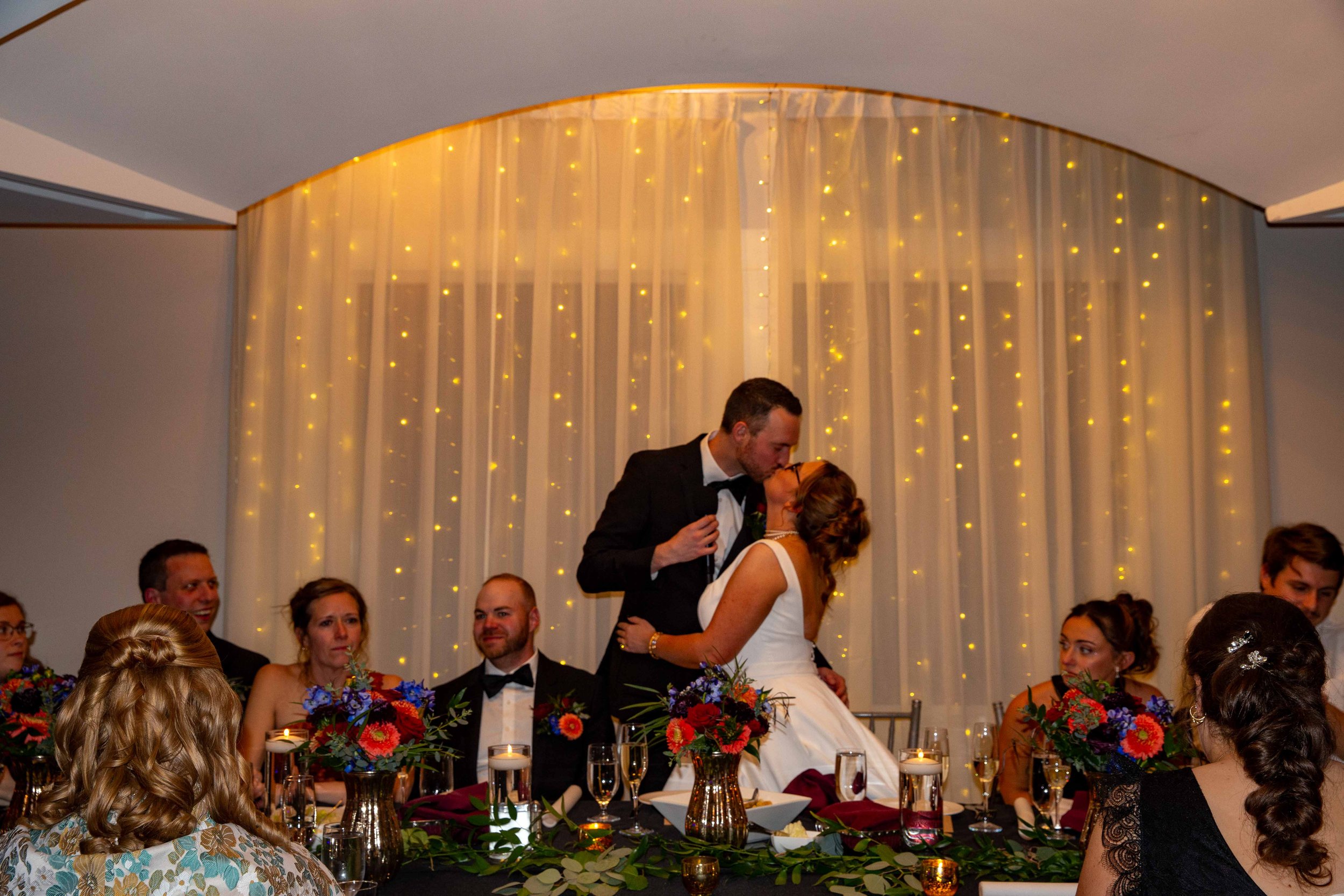 A newlywed couple sharing a kiss at their wedding reception while seated at a decorated table with flowers and candles. The background features a curtain with string lights, and other guests are seated around the table.