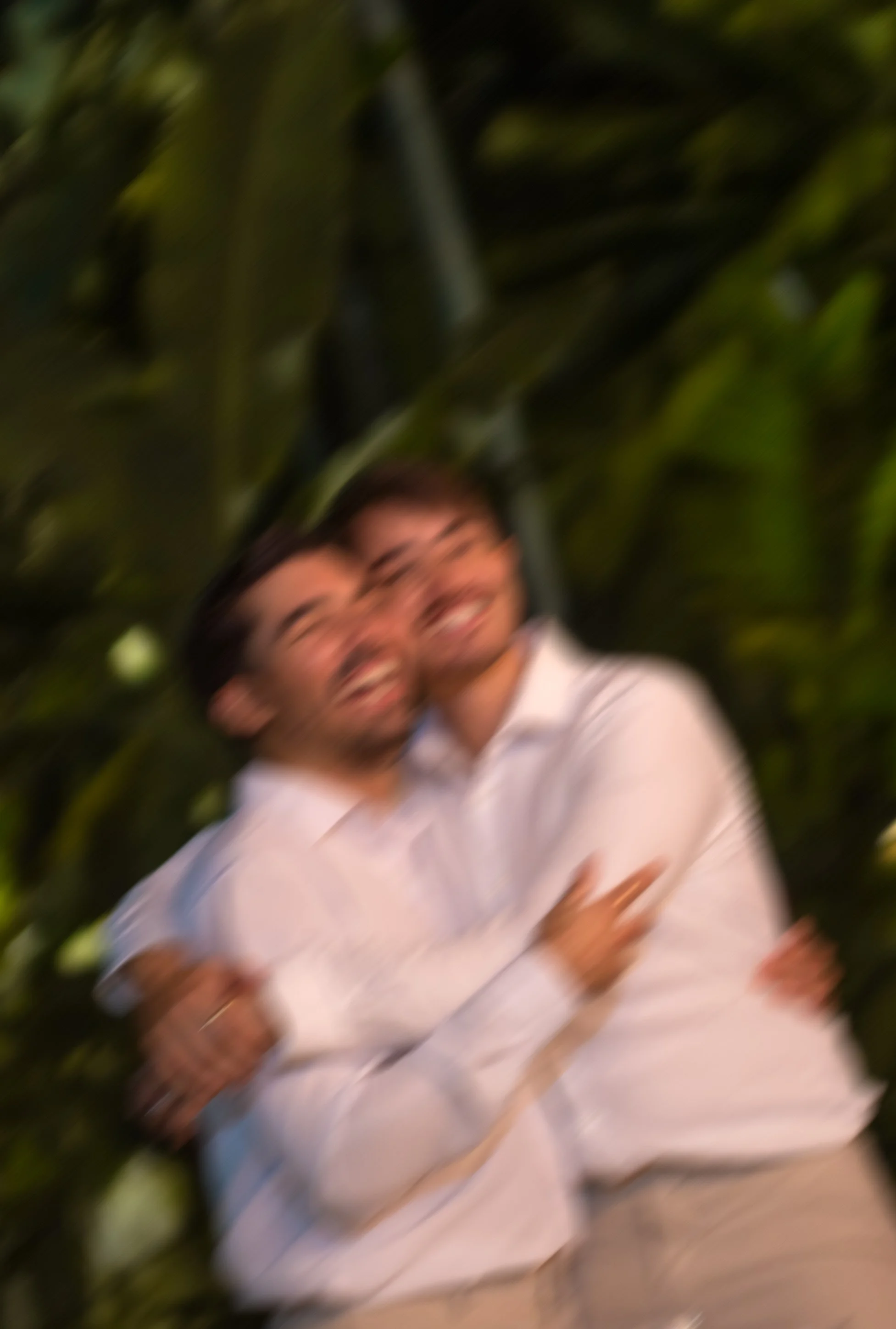 Two people smiling and hugging in front of green foliage.