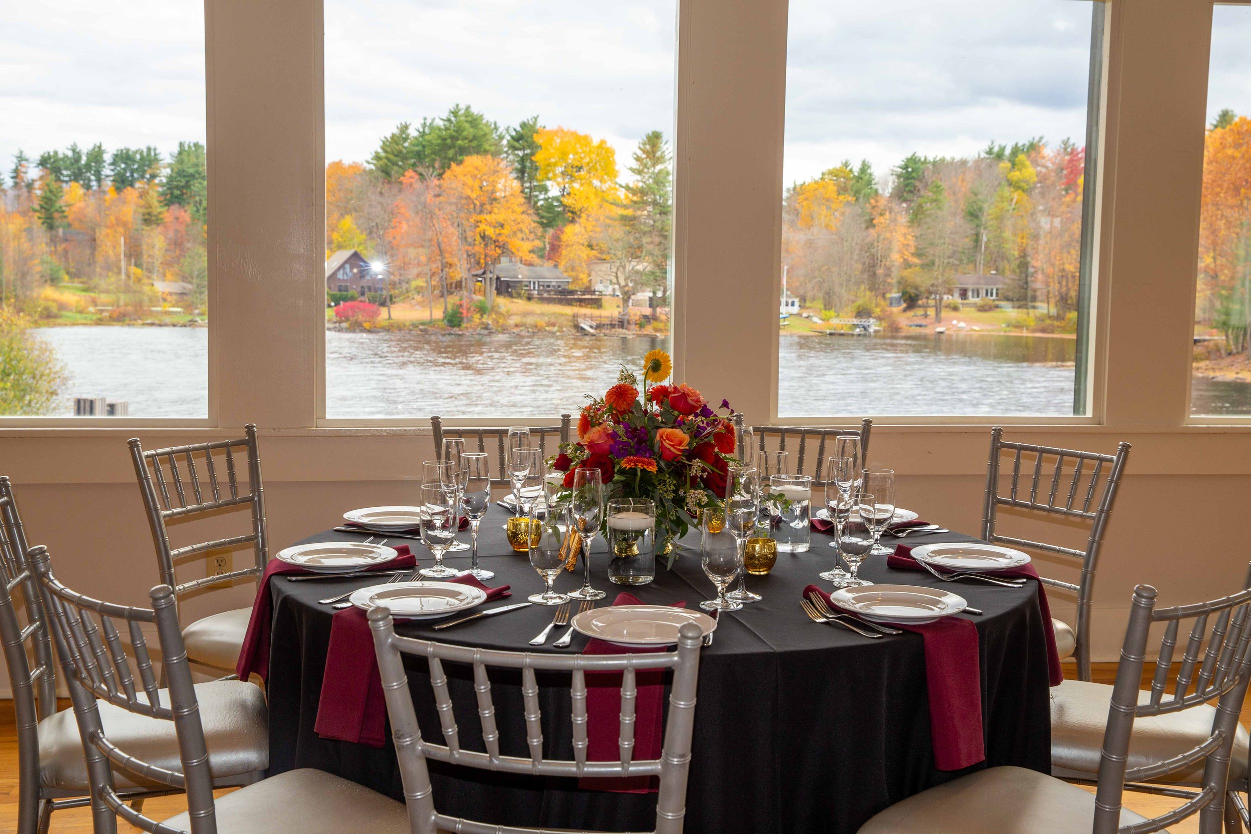 Round dining table set for a formal meal with a black tablecloth, surrounded by silver chairs, with a large floral centerpiece, and set with plates, silverware, glasses, and burgundy napkins, in front of large windows overlooking a lakeside with tree