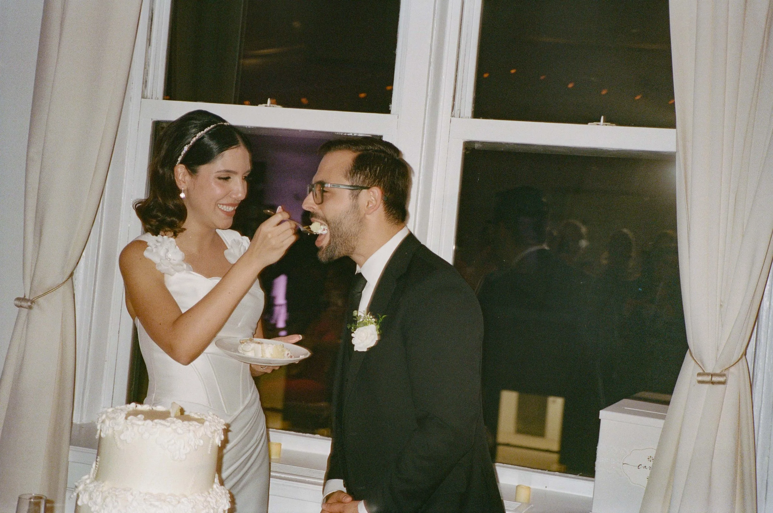 A bride feeds a slice of cake to a groom at a wedding reception. The bride is wearing a white dress and the groom is wearing a black tuxedo with a boutonniere. There is a decorated wedding cake on the table.