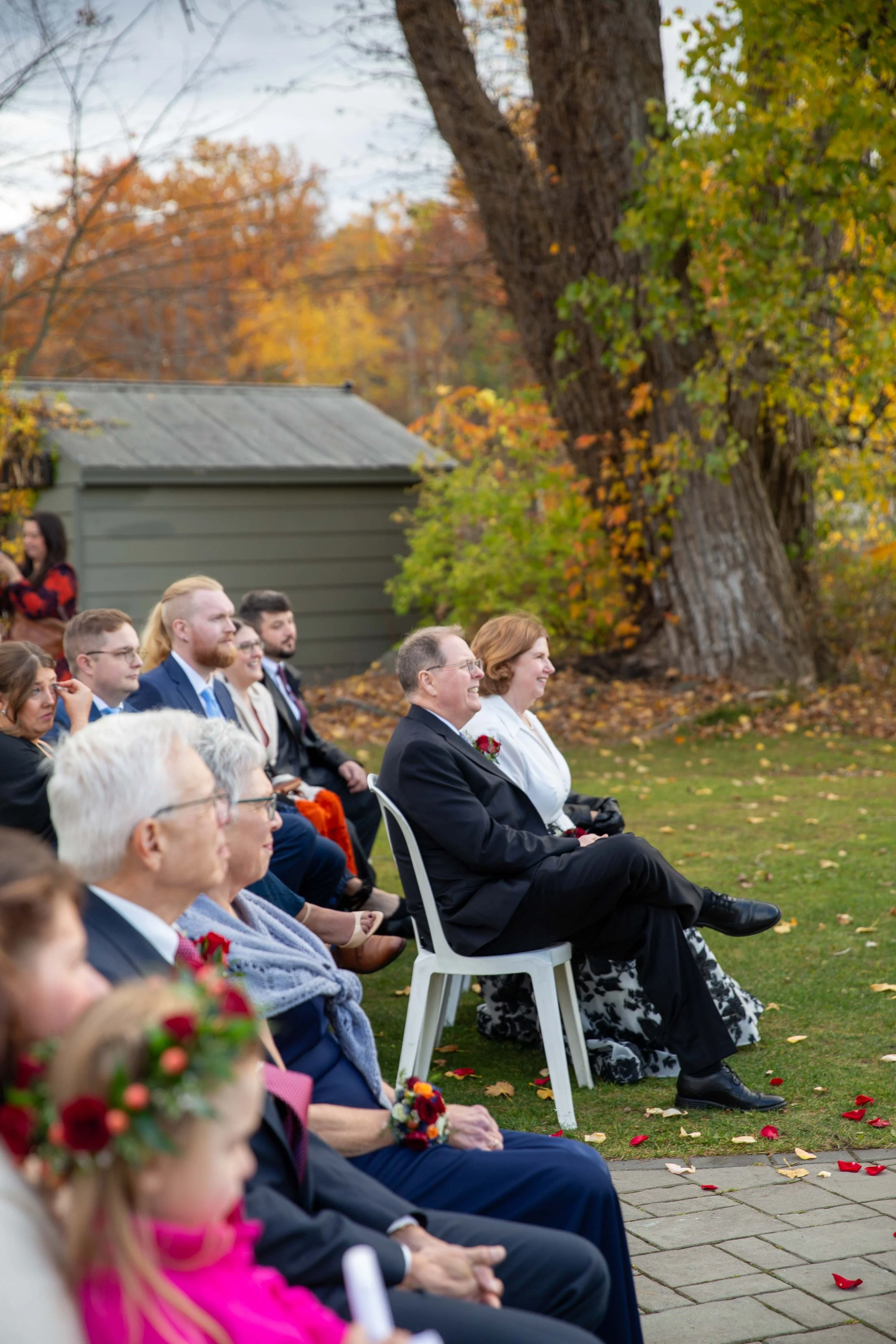 People seated outdoors during a wedding ceremony, with fall foliage in the background.