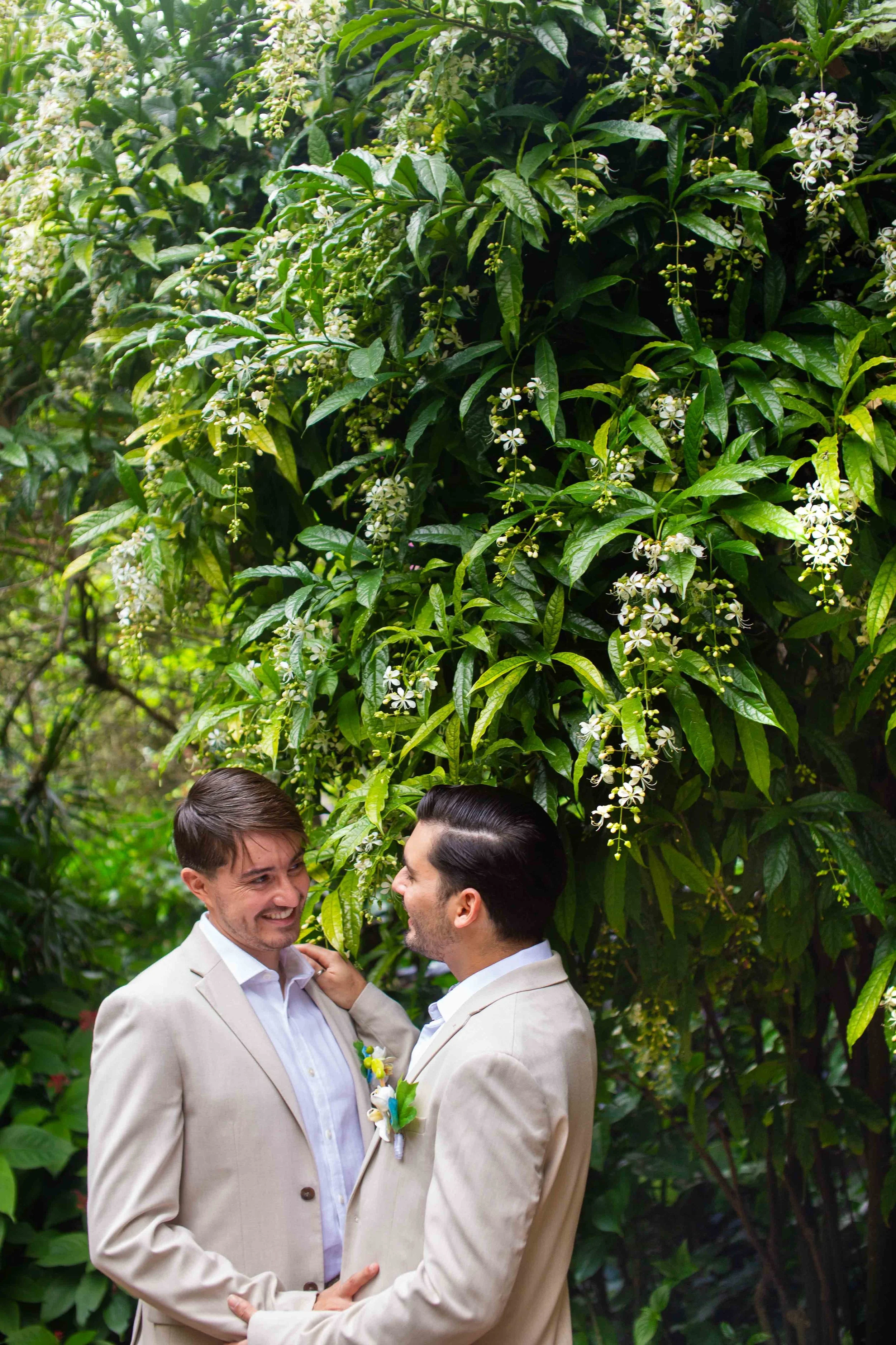 Two men dressed in cream suits share a tender moment, gazing into each other's eyes, surrounded by lush green foliage and white flowers, outdoors in a tropical setting.