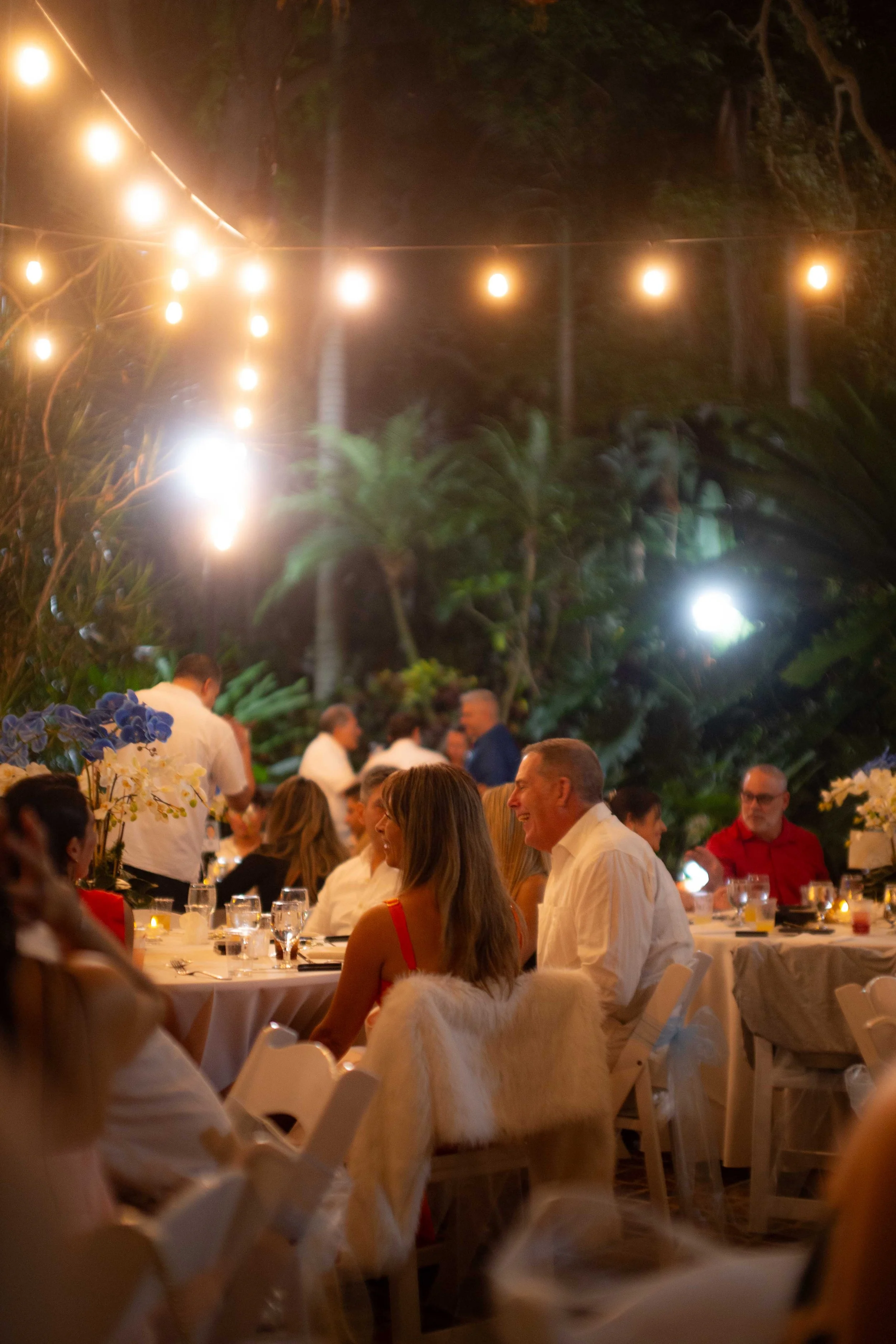 People dining at an outdoor event under string lights at night, with tropical trees in the background.