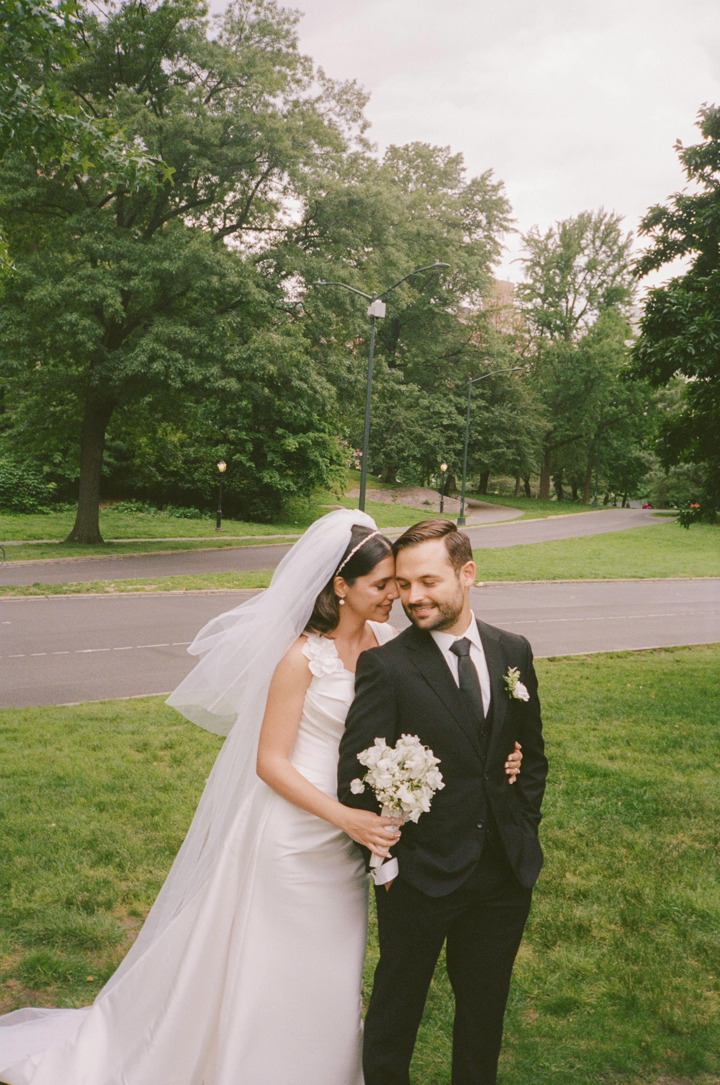 A bride and groom in wedding attire sharing an intimate moment, standing on a green lawn in a park with trees and a pathway in the background.