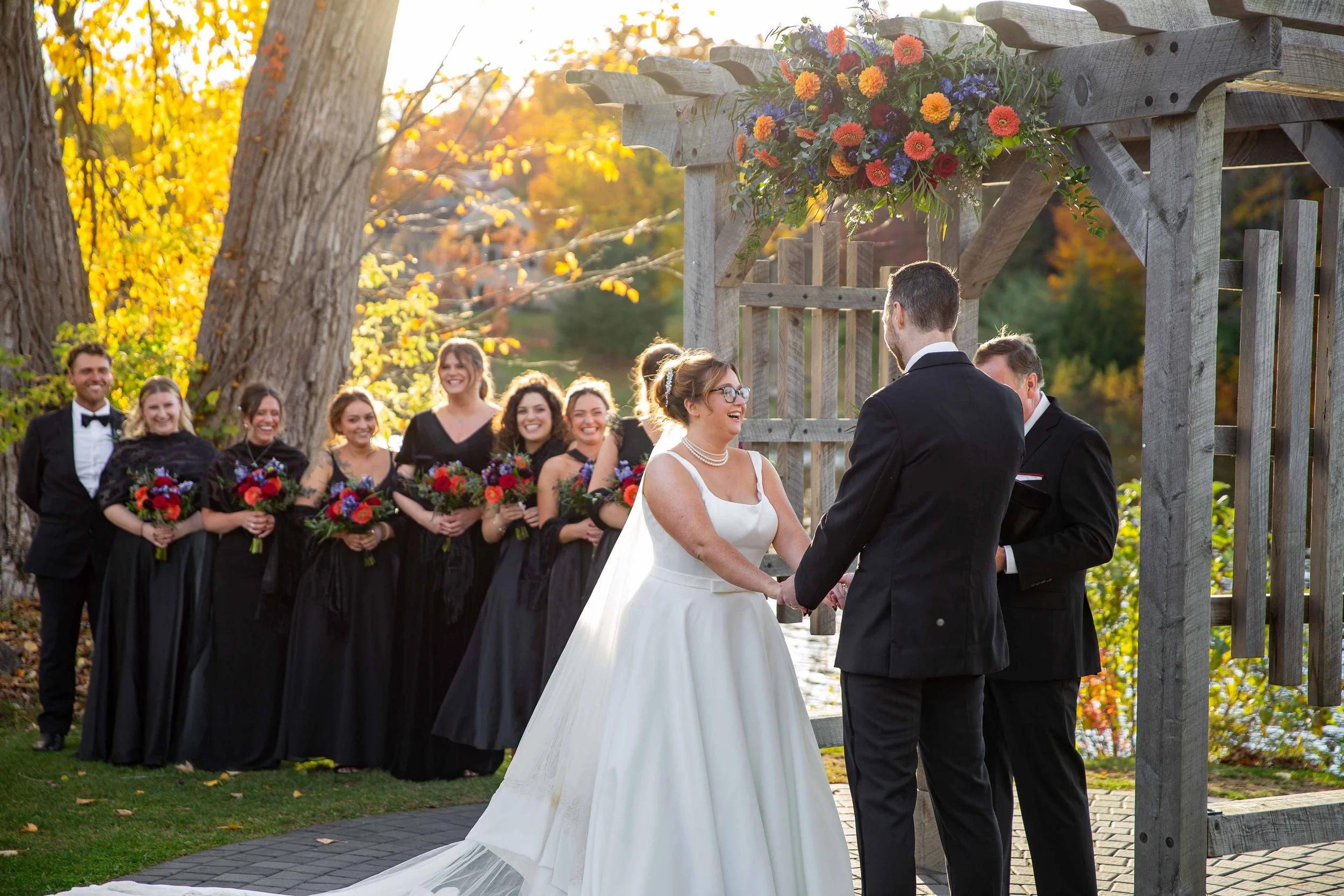 Bride and groom holding hands during their outdoor wedding ceremony, with the wedding party and guests smiling in the background, surrounded by trees with autumn foliage.