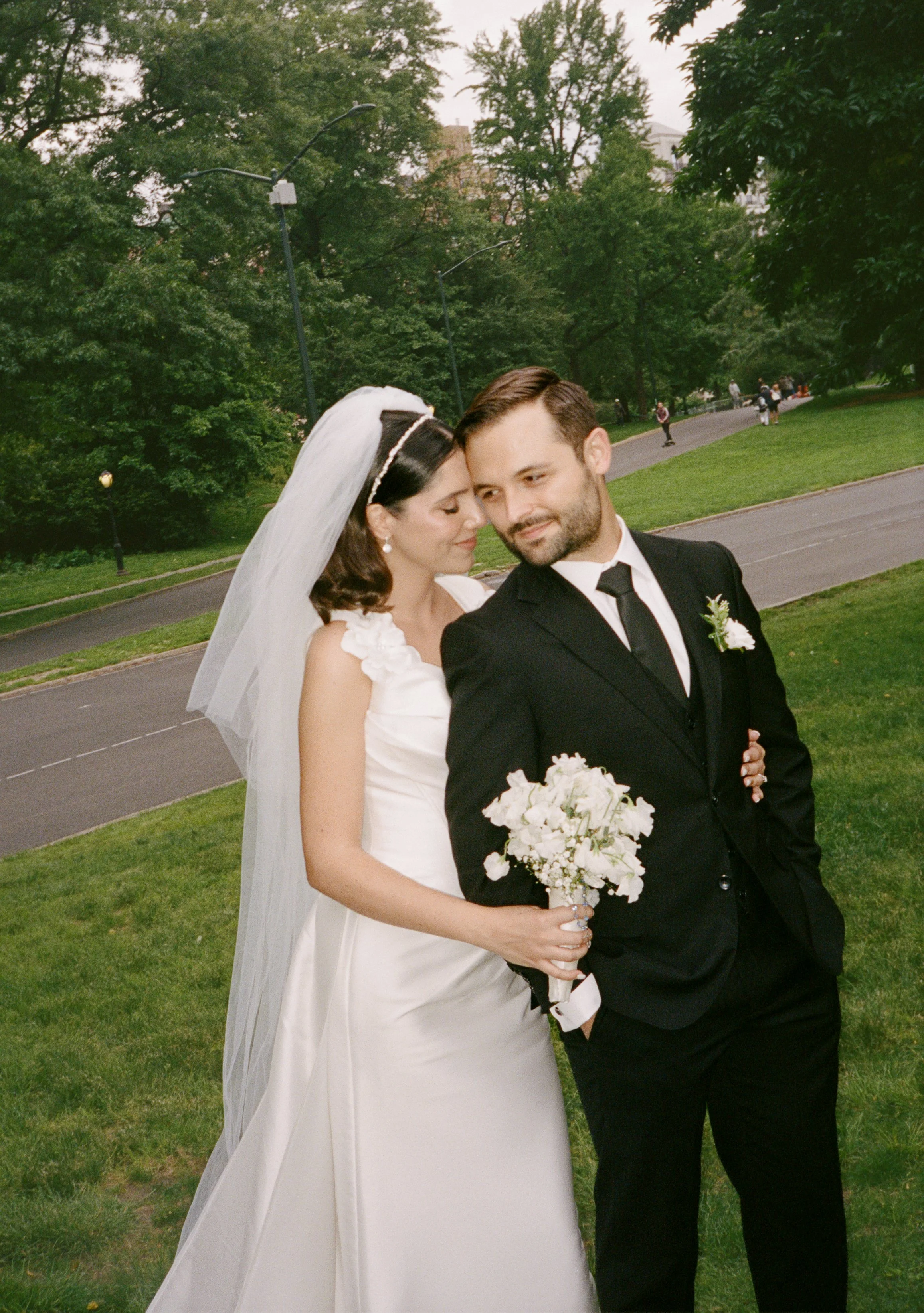 Bride and groom on their wedding day in a park, with the bride holding a bouquet of white flowers, both smiling and close to each other, surrounded by green trees and a few people in the background.