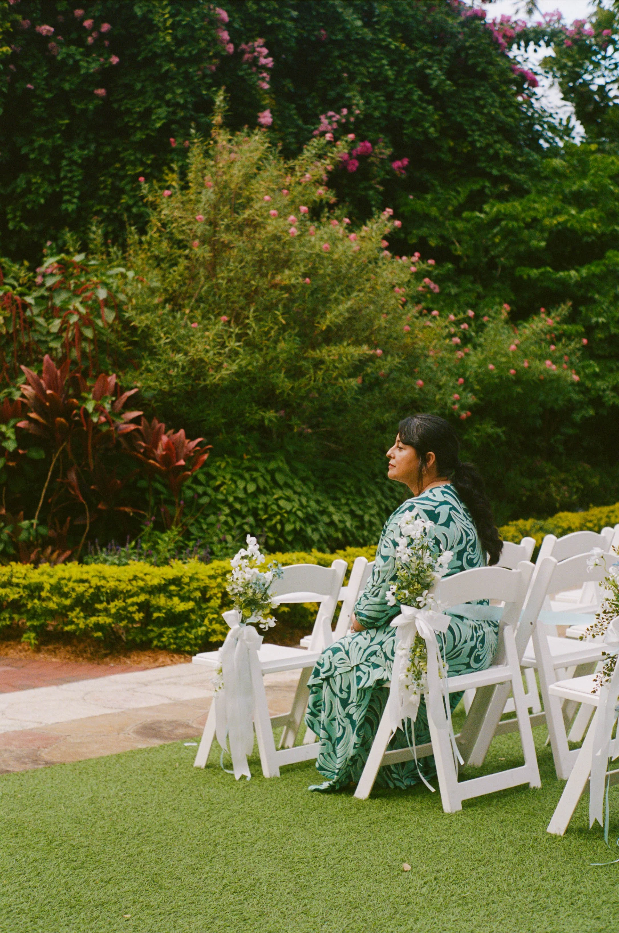 Woman sitting on white chairs decorated with flowers at an outdoor event, lush garden with pink flowers and green foliage in the background.