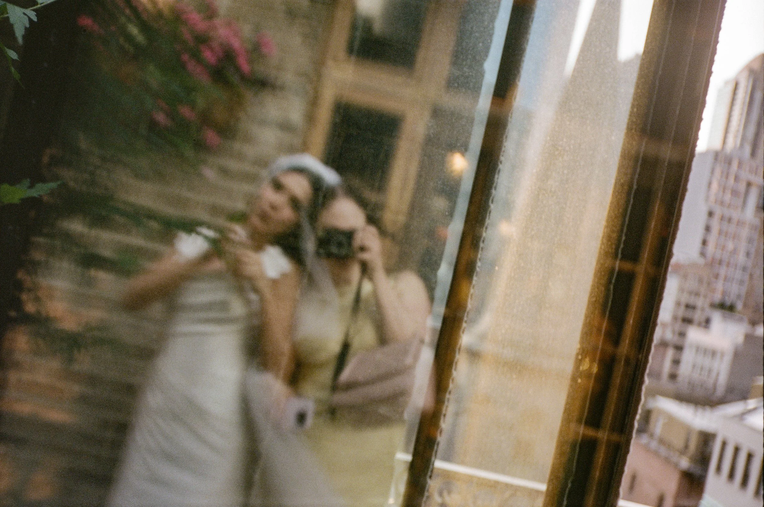A reflection of three women standing near a window on a city street, with buildings in the background, seen through a rain-speckled window.