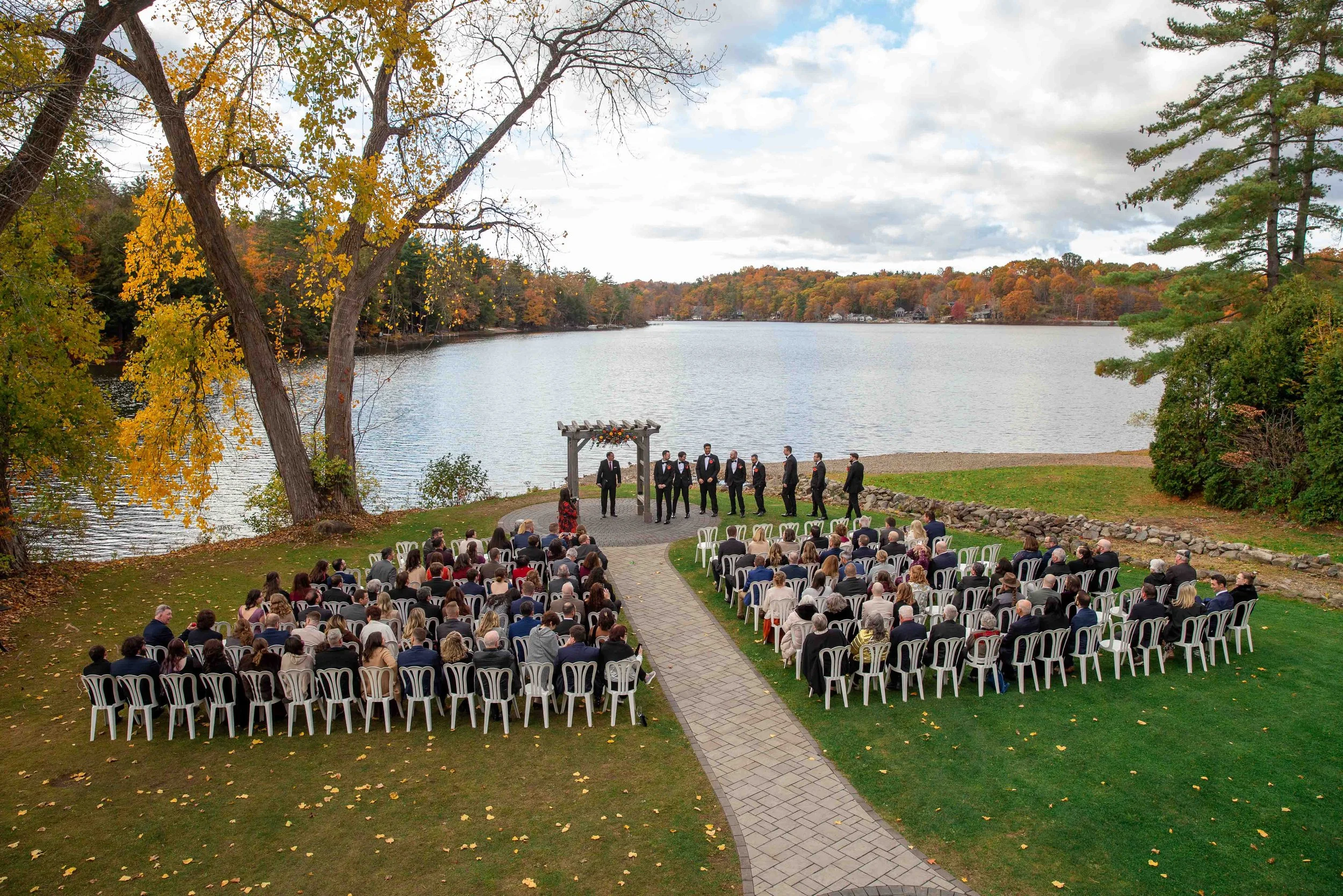 A wedding ceremony taking place outdoors by a lake during fall, with guests seated on white chairs facing a group of groomsmen standing under a wooden arch.