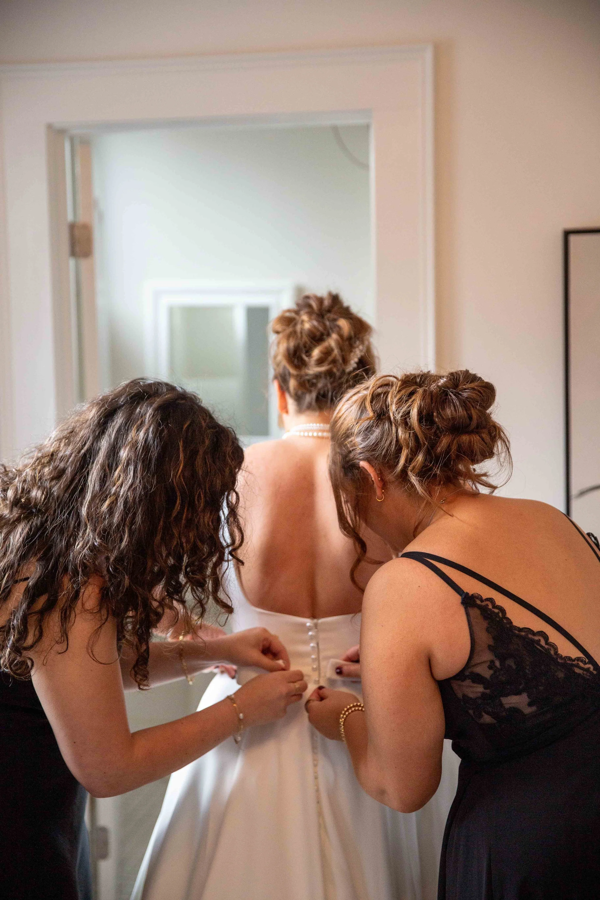 Two women helping a bride button up the back of her white wedding dress.