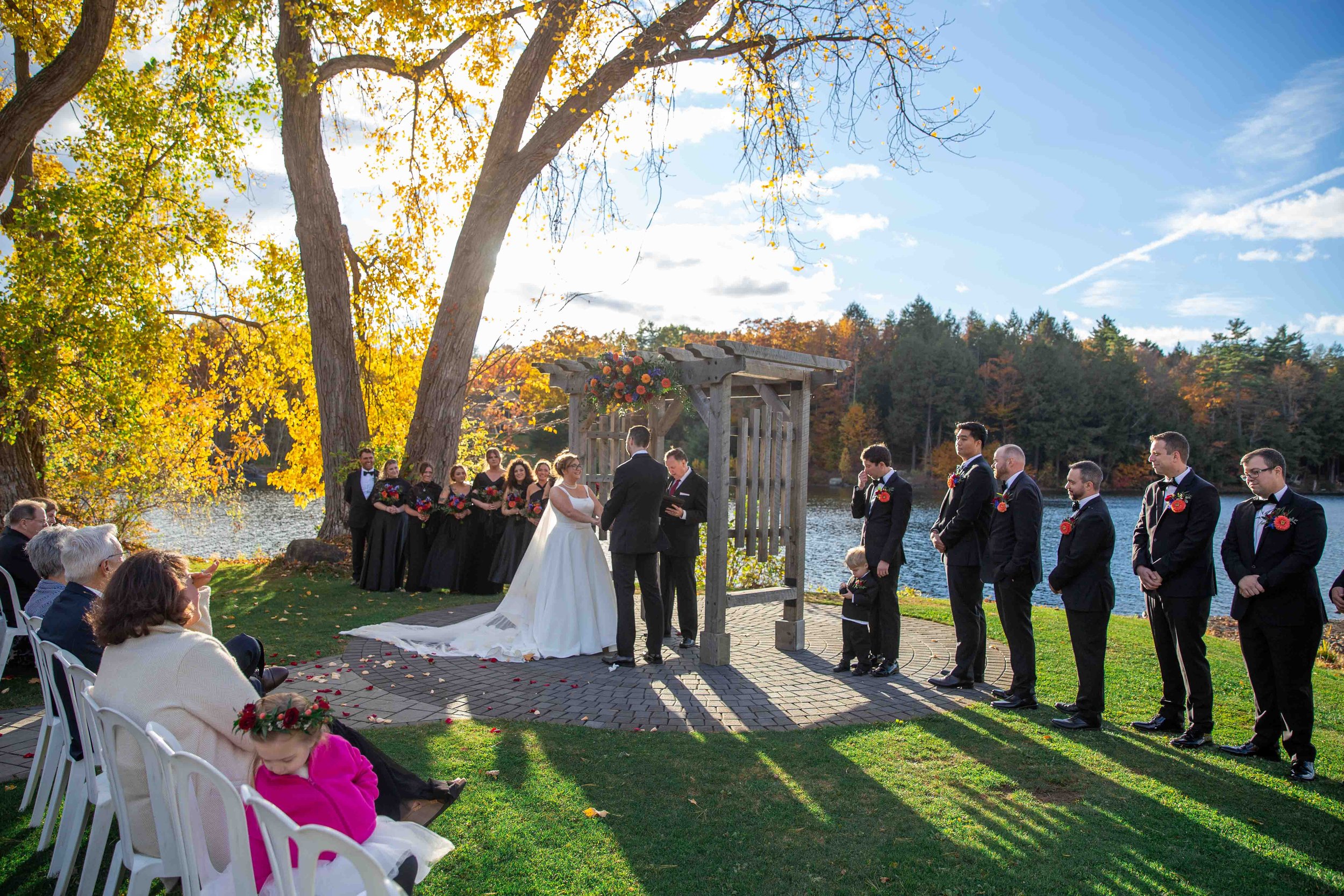 Outdoor wedding ceremony by a lake on a sunny autumn day, with a bride and groom exchanging vows under a wooden arch decorated with flowers, surrounded by bridesmaids, groomsmen, and seated guests.